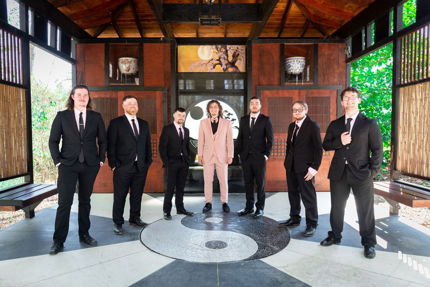 A group of seven people in formal attire stand together in a covered outdoor walkway with wooden beams and walls.
