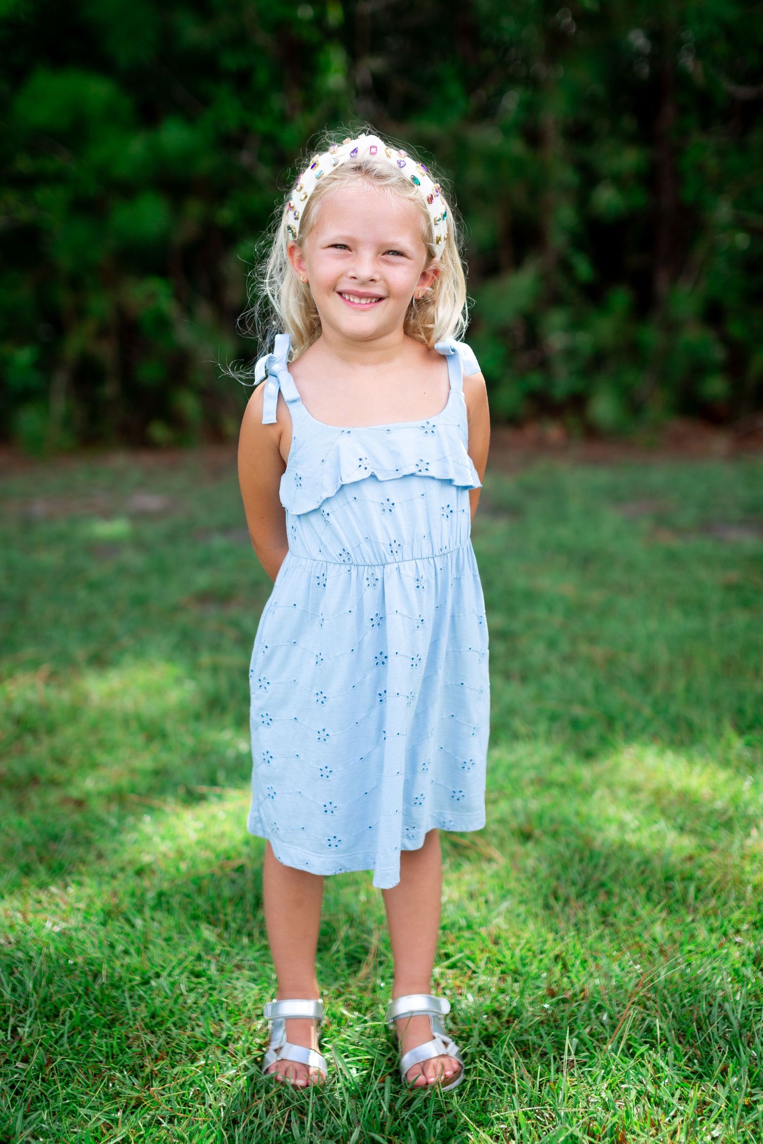 Young child in a blue dress playing and smiling on green grass in a garden setting.