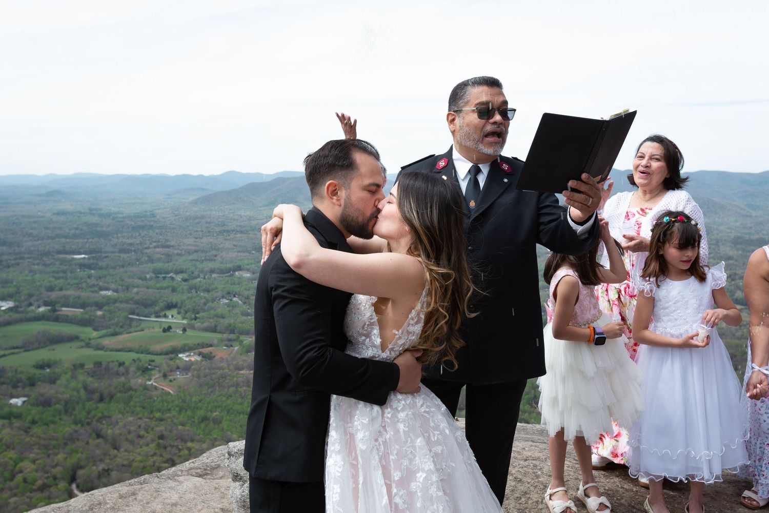 Couple shares first kiss at mountain top wedding ceremony with officiant and flower girls in white dresses.