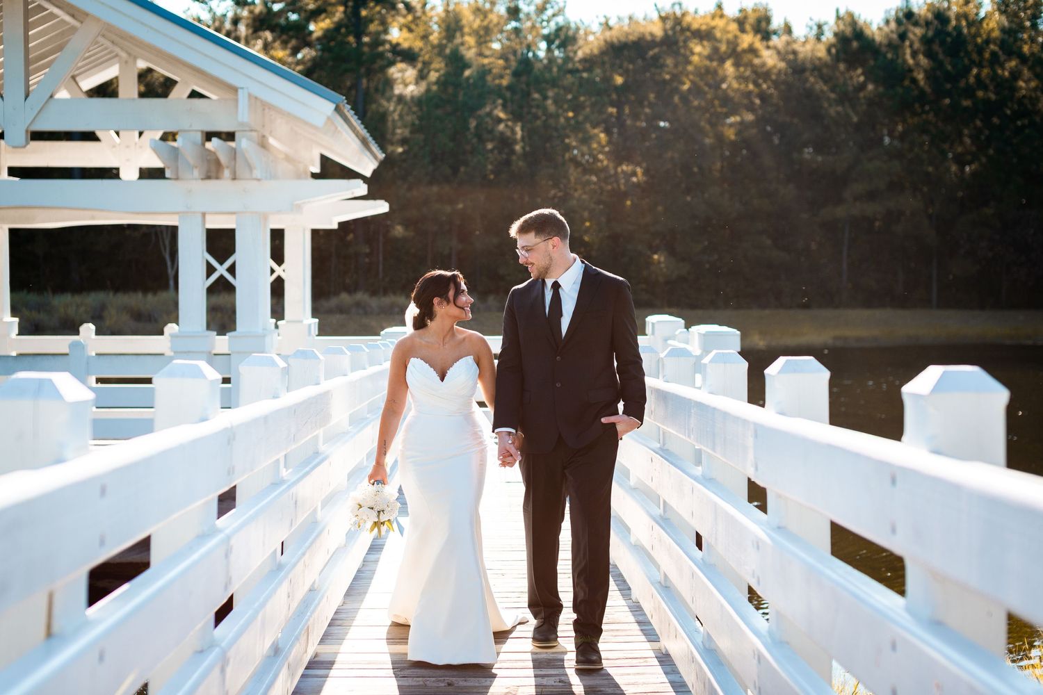 Wedding couple walks hand in hand on a white wooden deck surrounded by nature.