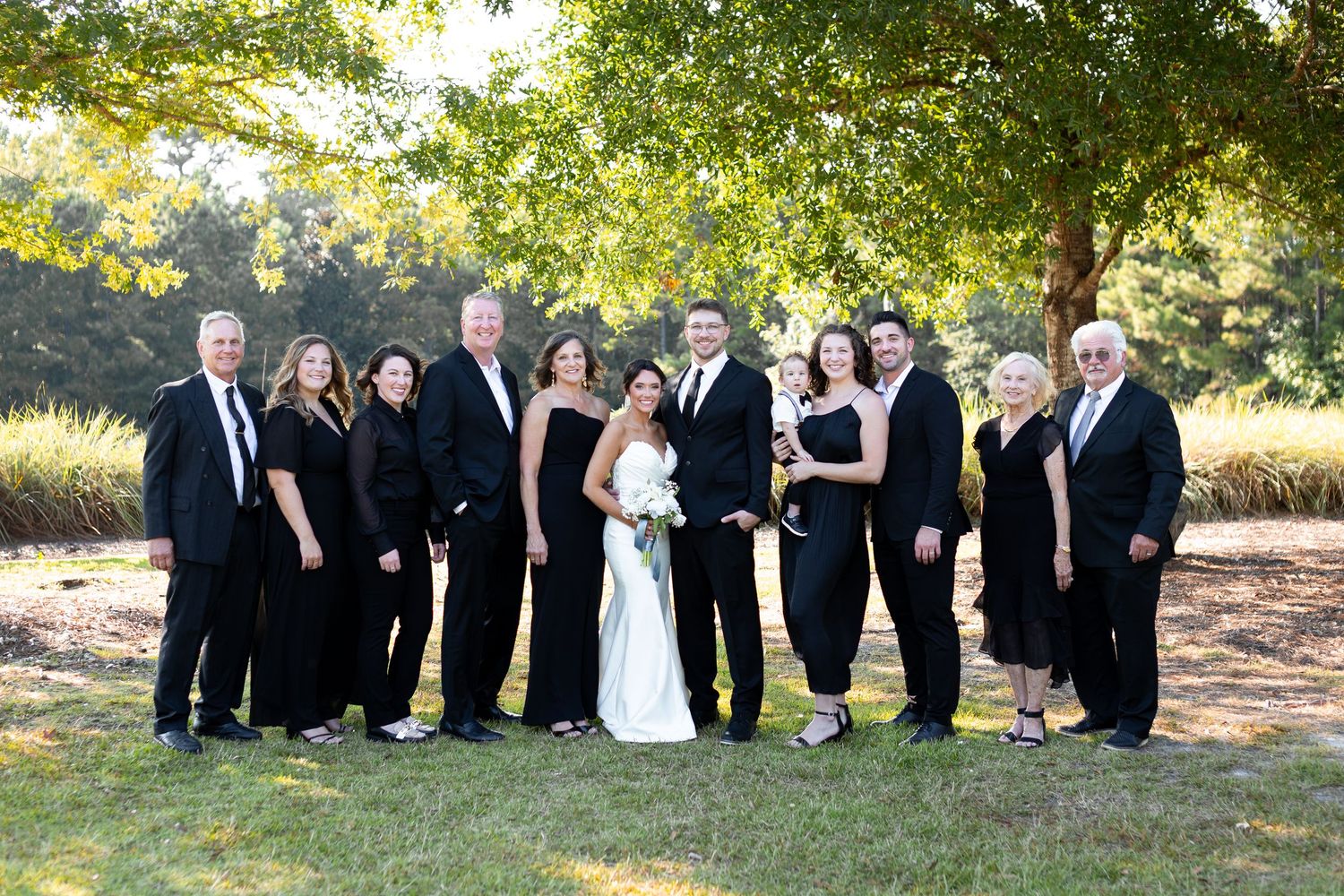 Wedding party in formal black attire poses together under trees during an outdoor celebration.