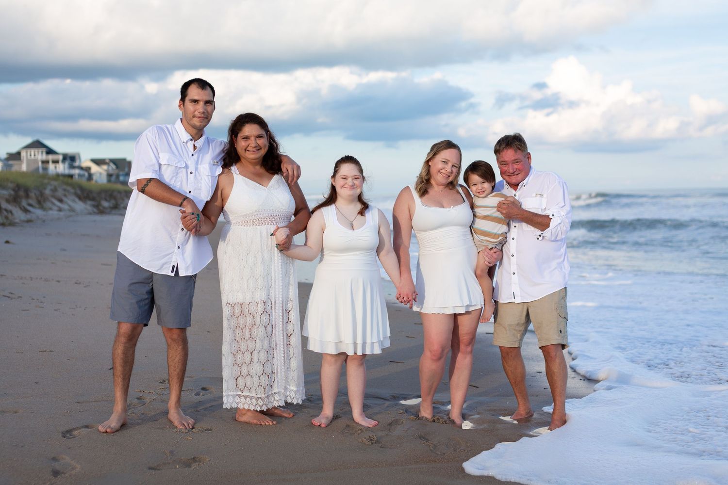 Family group poses together in white outfits for casual beach photos at sunset.