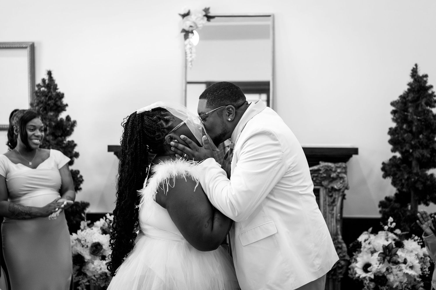 Black and white photo of a couple sharing their first kiss at a wedding ceremony with a bridesmaid watching nearby.