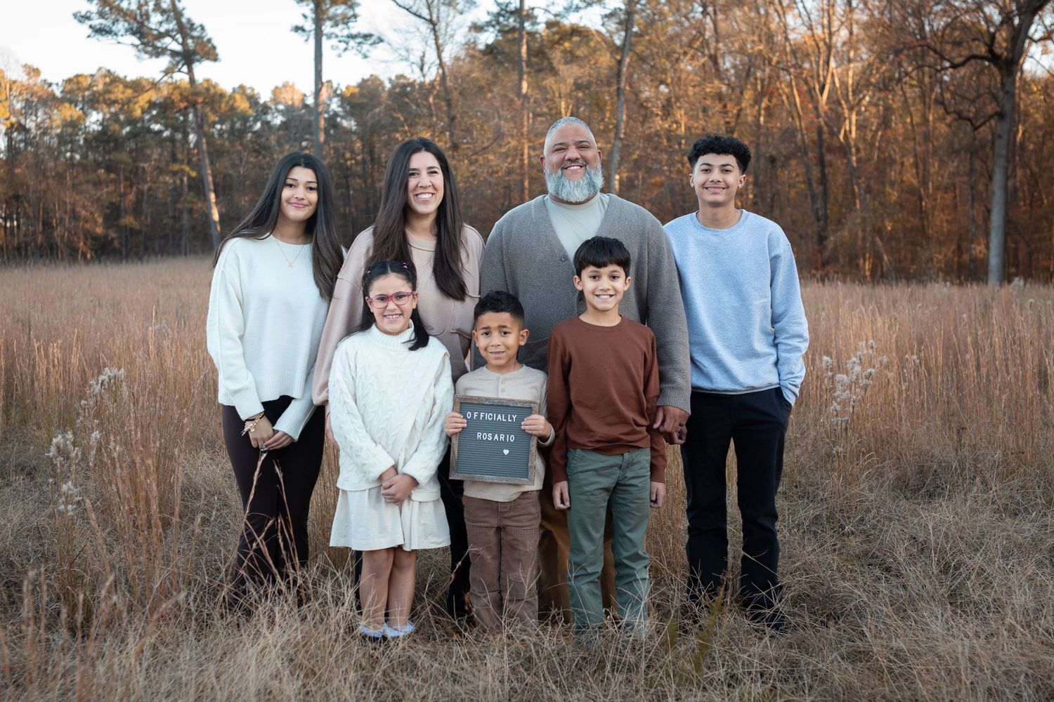 Family of seven posing outdoors in a golden field surrounded by autumn trees.