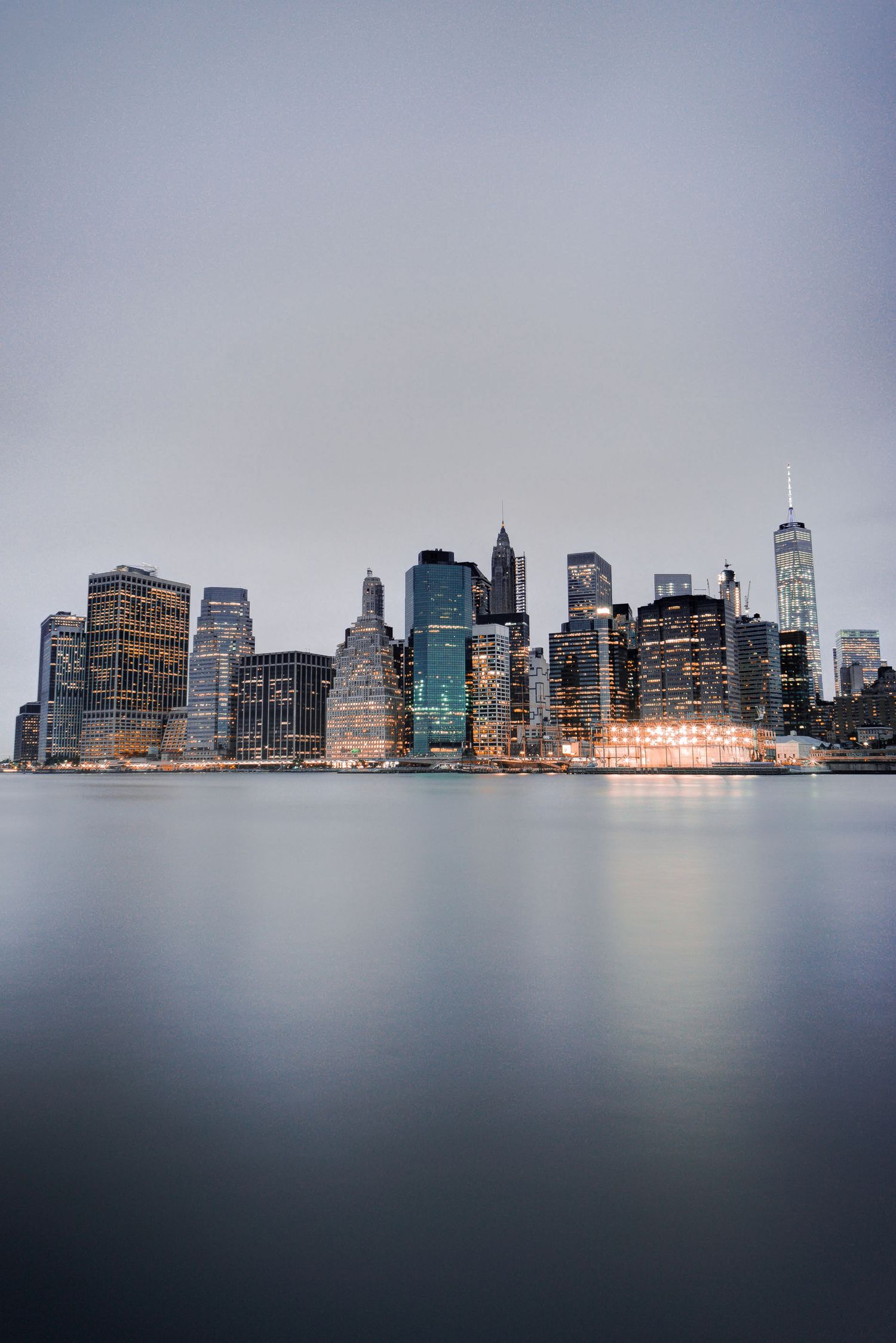 Manhattan skyline reflecting on calm water at dusk, featuring One World Trade Center and illuminated skyscrapers under overcast sky.