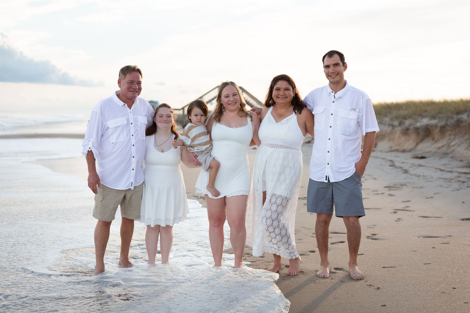 Family of six posing together on a sandy beach at sunset, wearing white outfits, smiling with ocean waves at their feet.