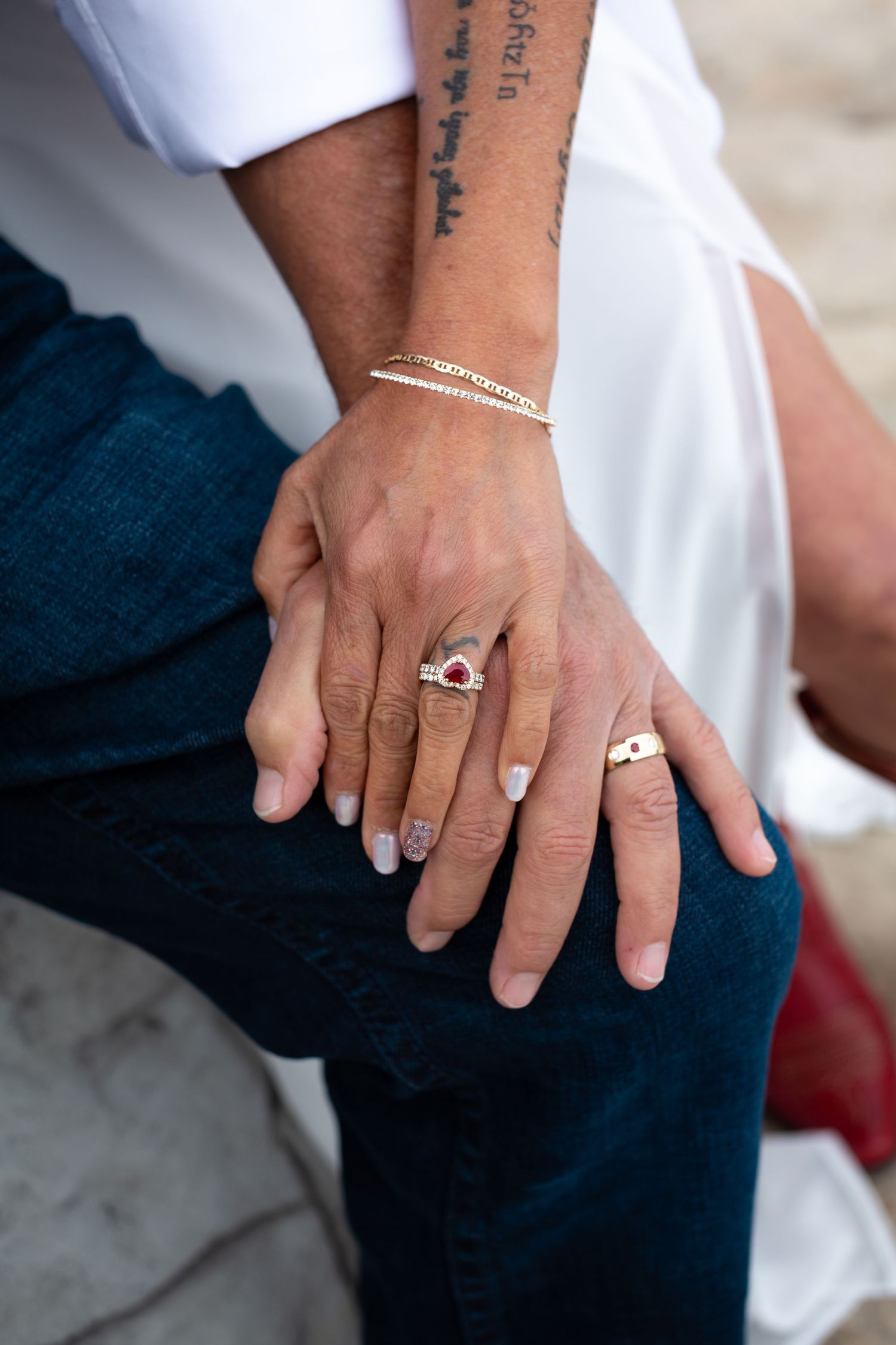 Couple holding hands showing engagement rings, bracelet, and tattoo, wearing white top and blue jeans.
