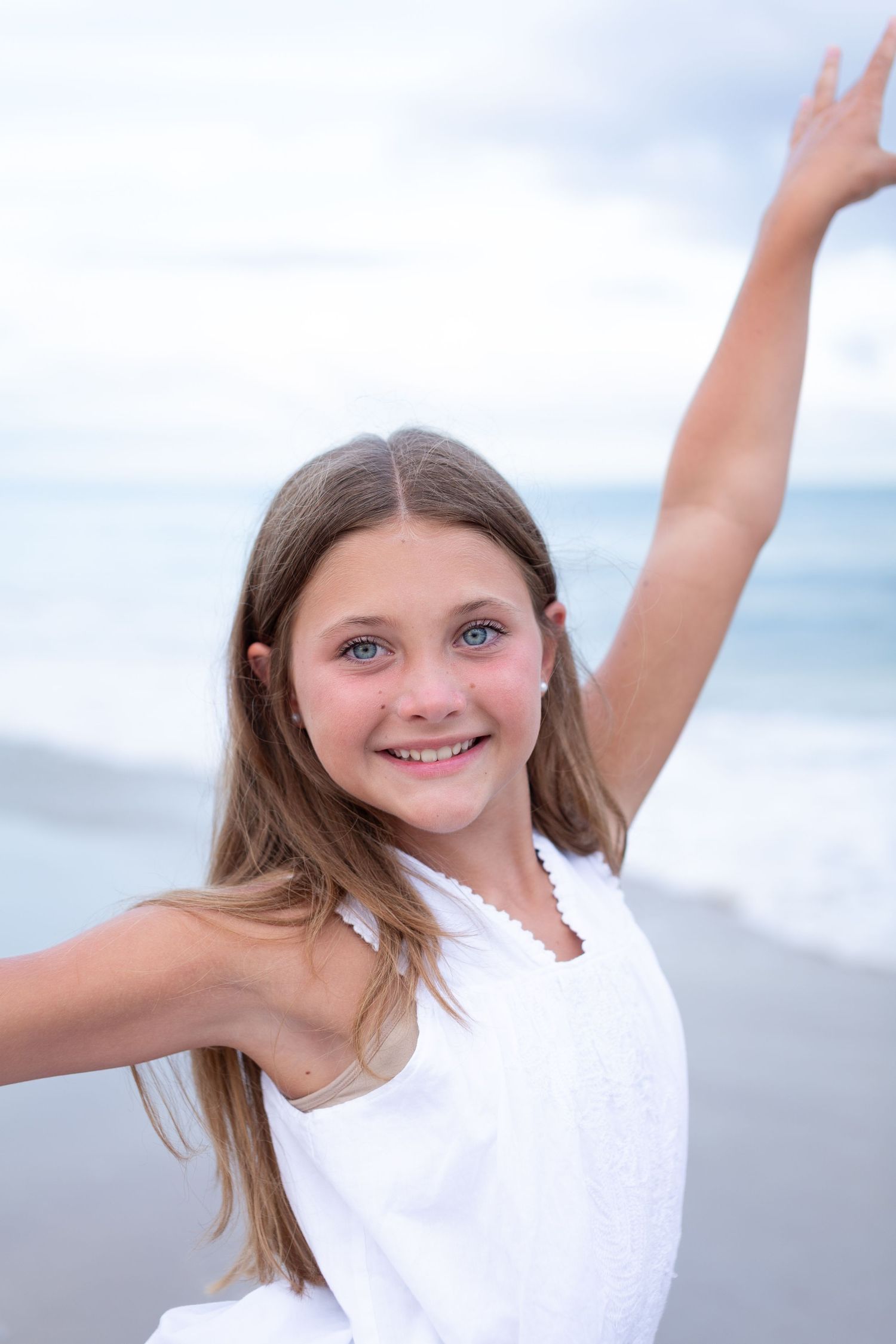 Young girl in white dress joyfully poses with arms raised on a beach at sunset.