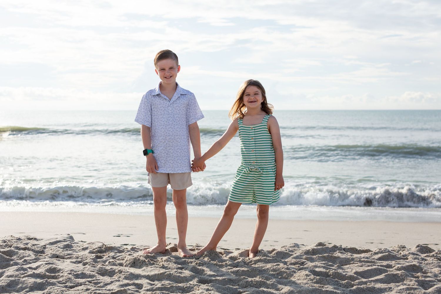 Two young people holding hands while standing on sunny beach in summer attire.