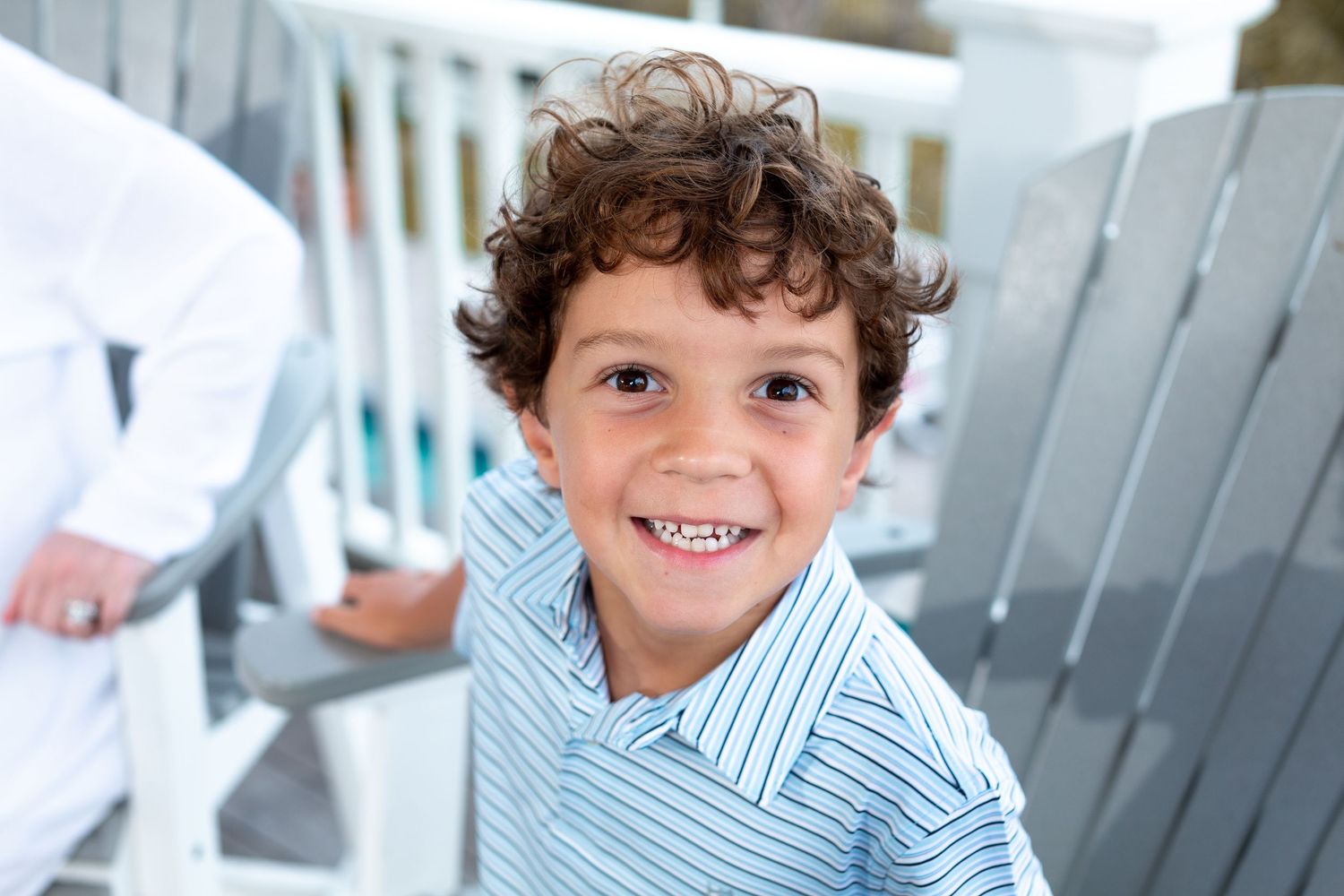 Young child wearing a blue striped shirt smiles brightly while sitting on a white porch chair.