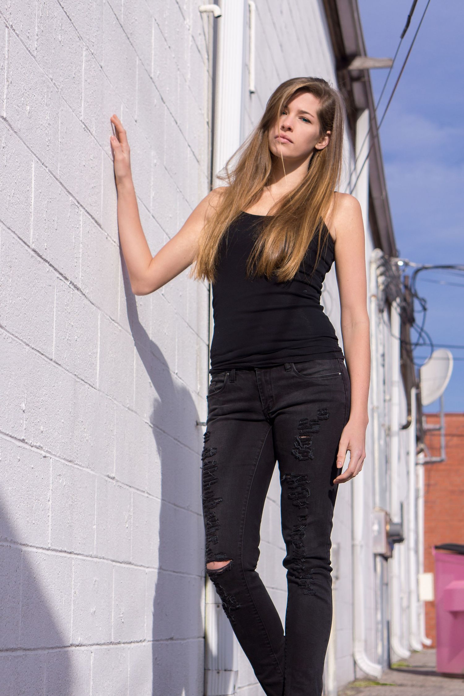 A person in black ripped jeans and tank top poses against a white wall with power lines visible in the background.