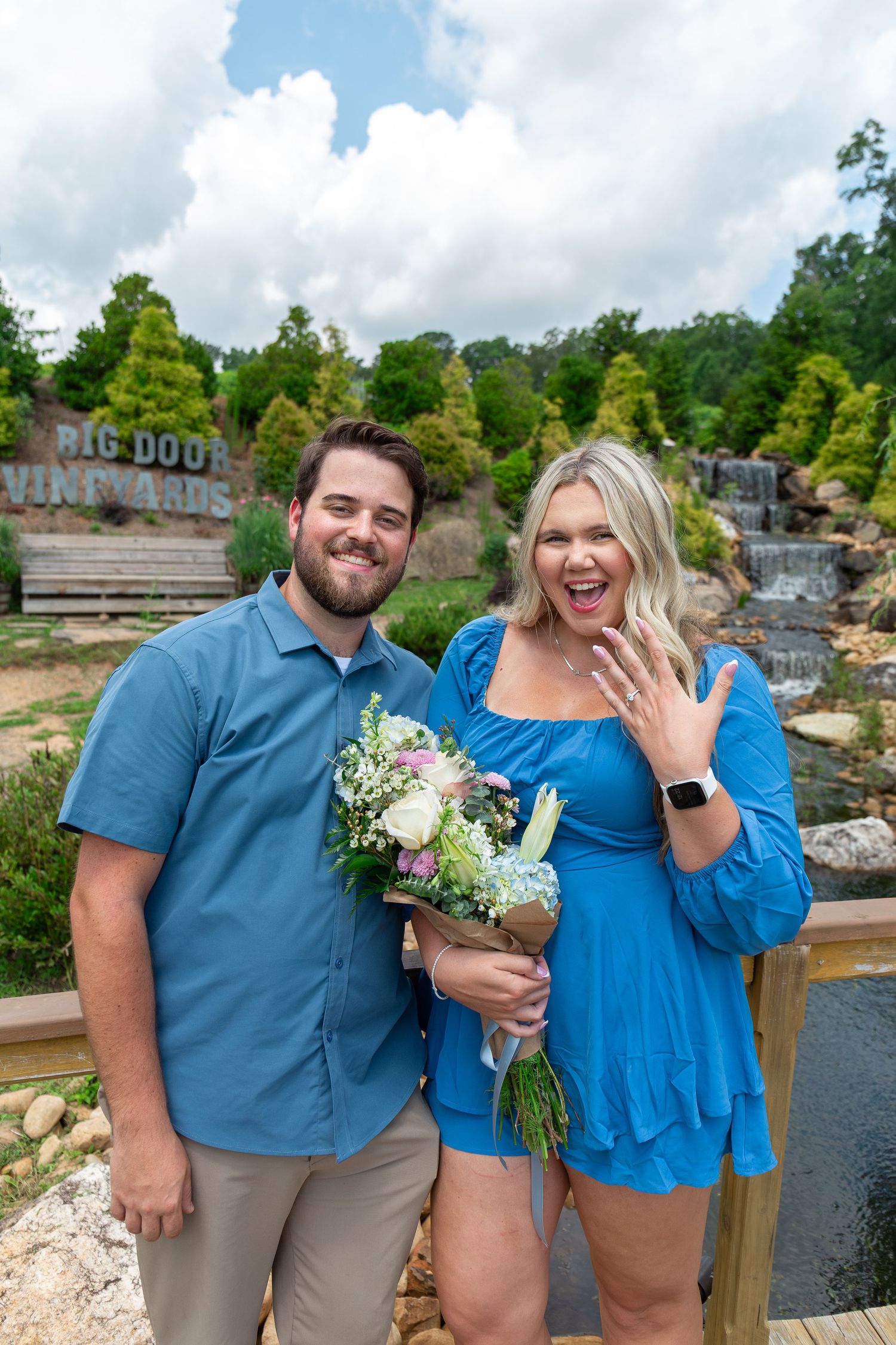 Couple celebrating engagement at Big Door Vineyards, woman in blue dress showing ring while holding bouquet.
