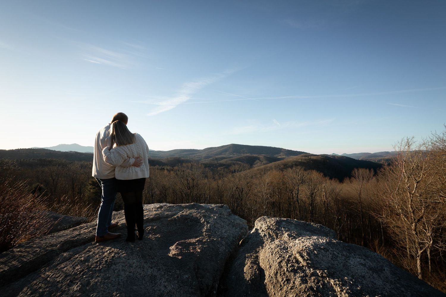 Hiker stands on rocky mountain overlook with expansive valley views on a clear sunny day.