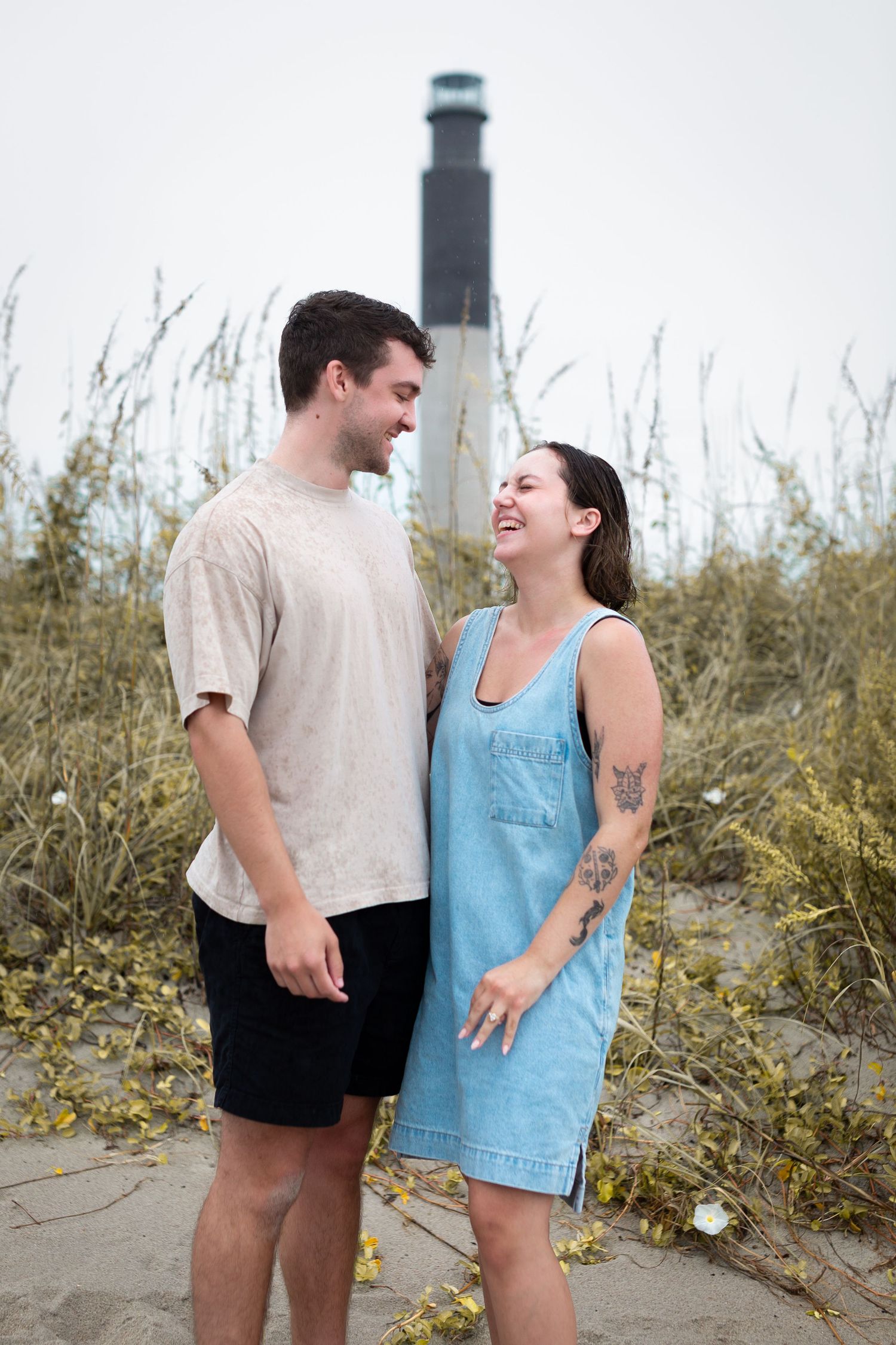 A couple shares joyful laughter while standing together on a beach with sea grass and dunes in the background.