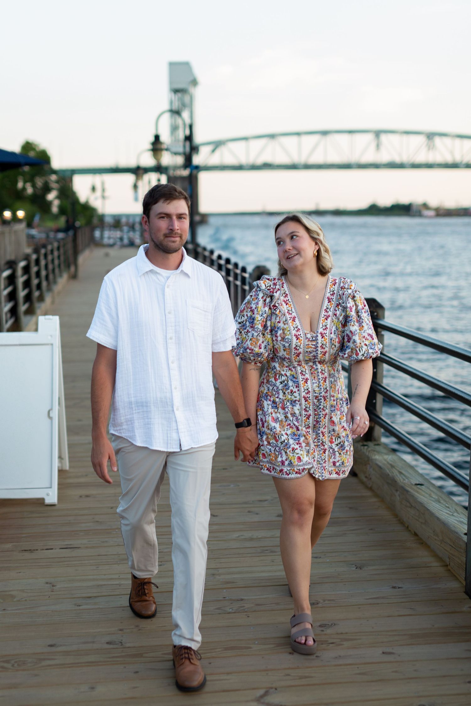 A couple walks together on a wooden boardwalk by the water during sunset, with a bridge visible in the background.