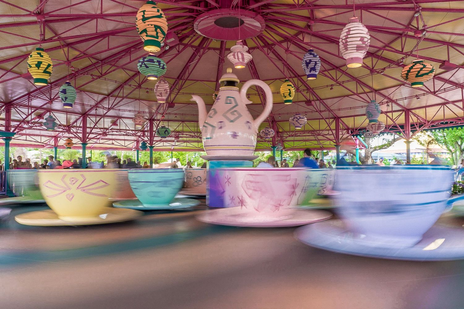 Colorful spinning teacup ride at a theme park with motion blur, featuring a large teapot centerpiece under a decorative canopy.