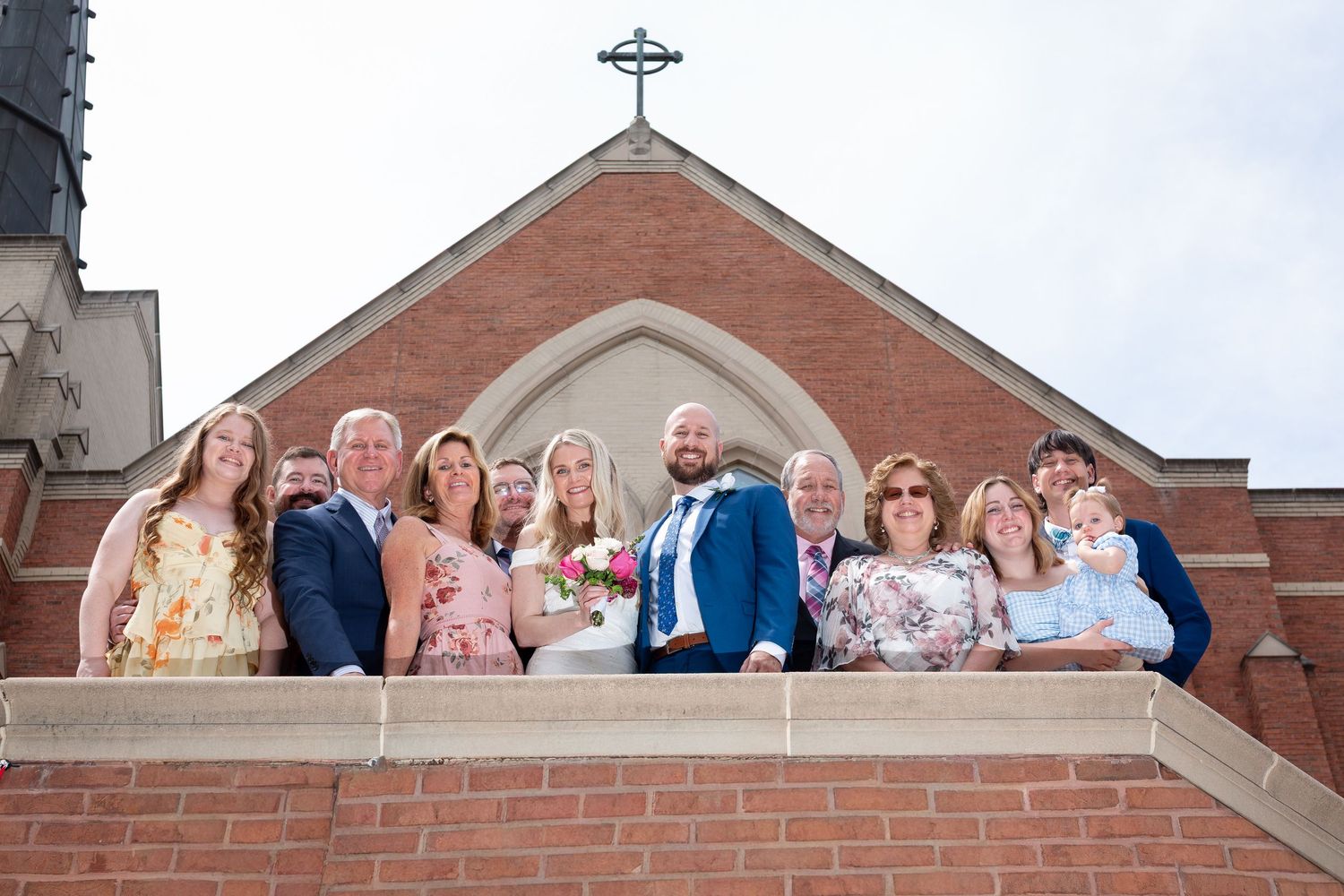 Wedding party poses on church steps in front of a brick building with a cross, celebrating a joyful ceremony.