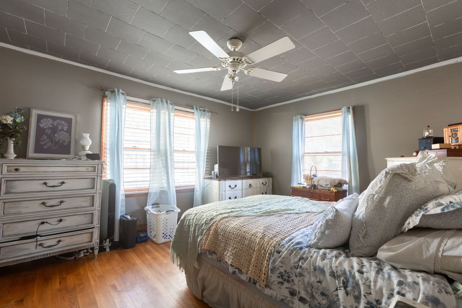 A cozy bedroom with gray walls, hardwood floors, and a ceiling fan features large windows letting in natural light.