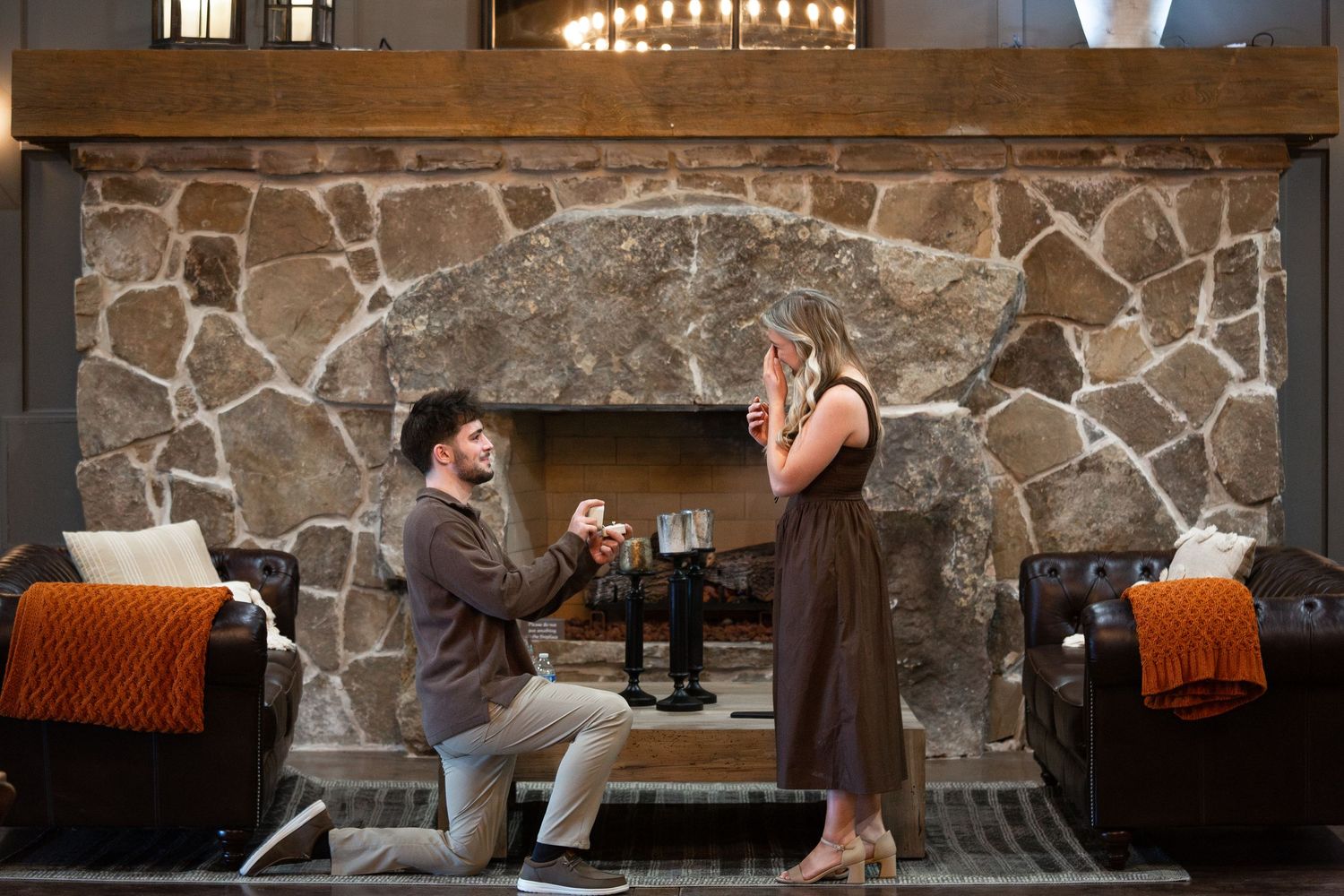A couple shares a special moment during a proposal in front of a grand stone fireplace with orange throw pillows on chairs.
