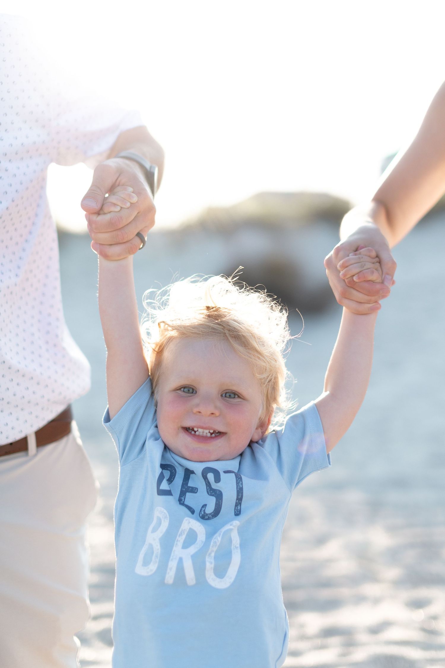 Young child wearing light blue 'Best Bro' t-shirt being lifted by hands on sunny beach.