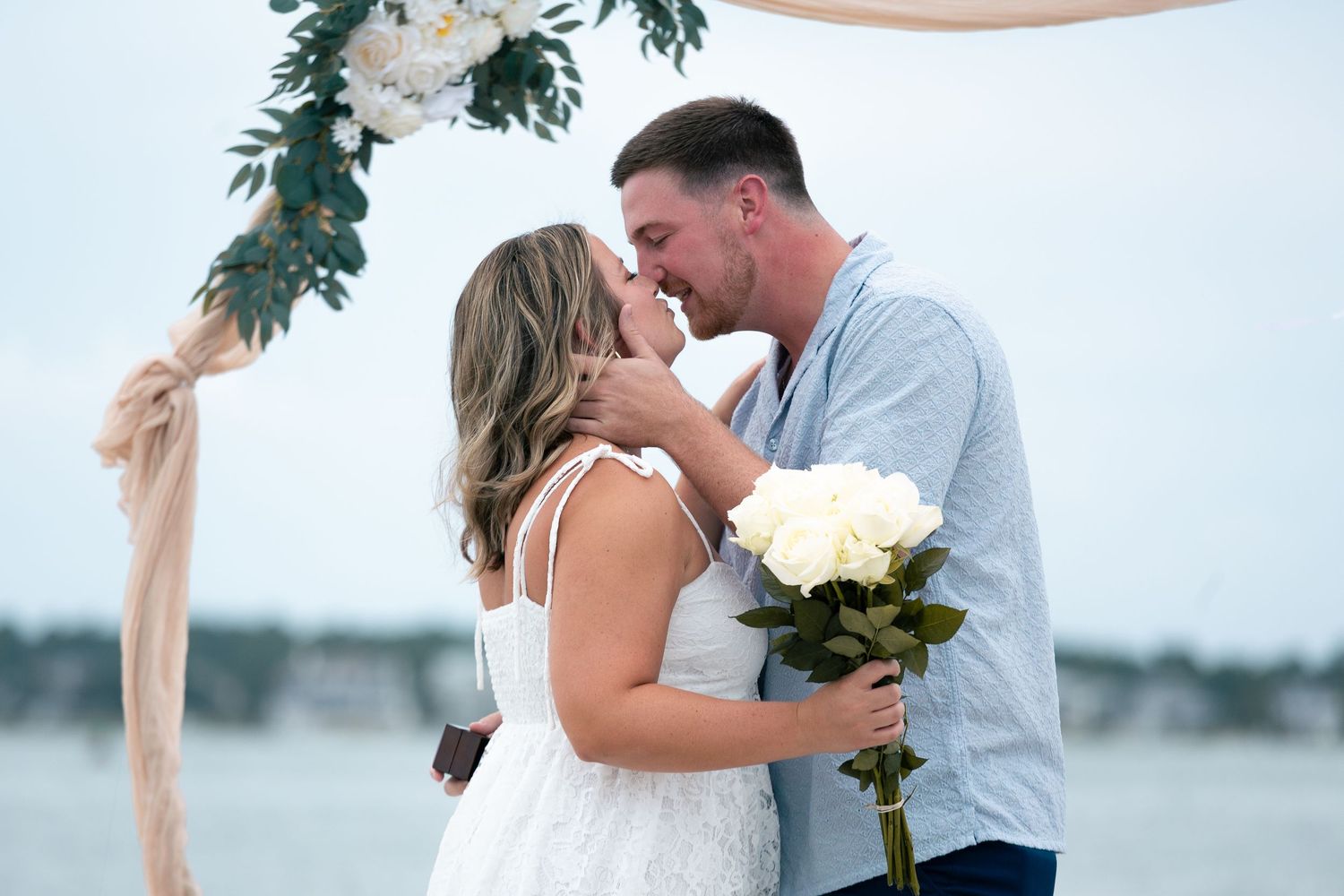 A couple embraces and kisses during their beach wedding ceremony with white flowers.