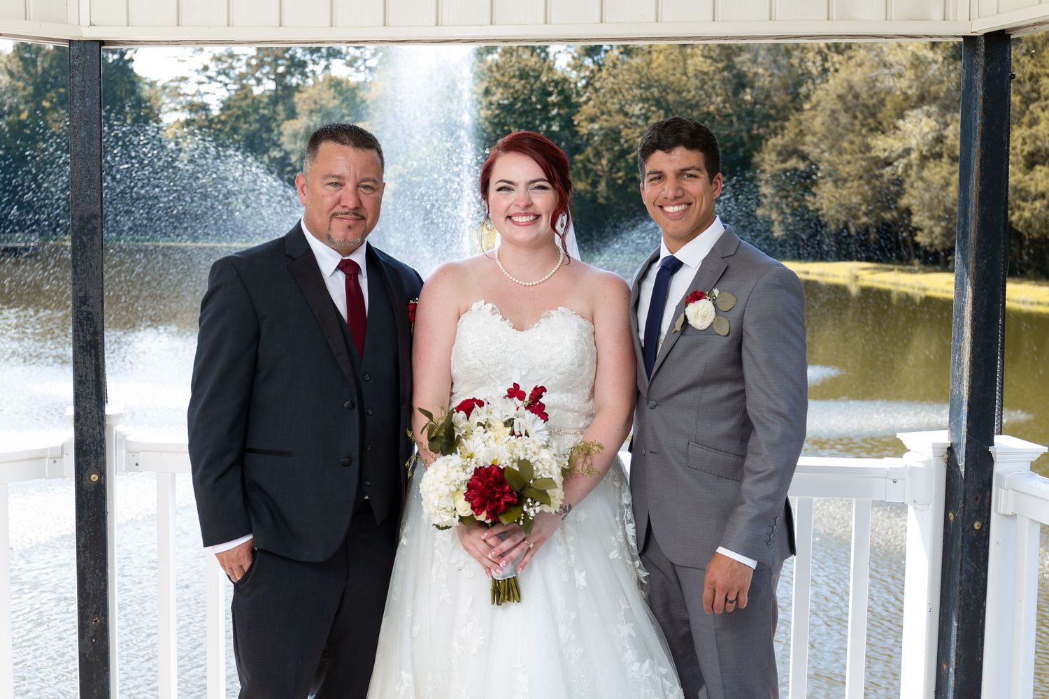 Wedding party poses together in formal attire near a lakeside gazebo.