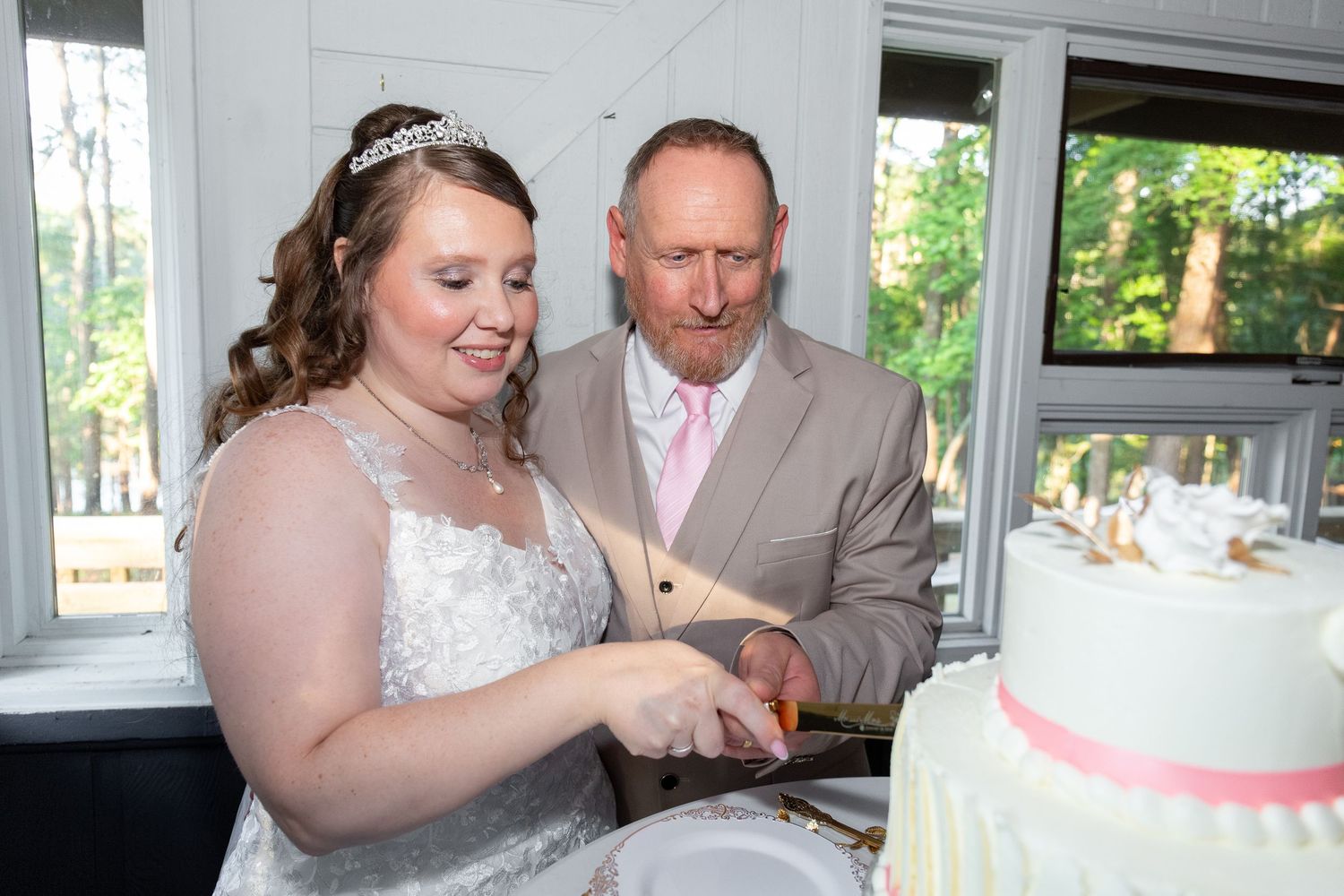 Bride and groom cutting their white wedding cake with pink ribbon at a reception.