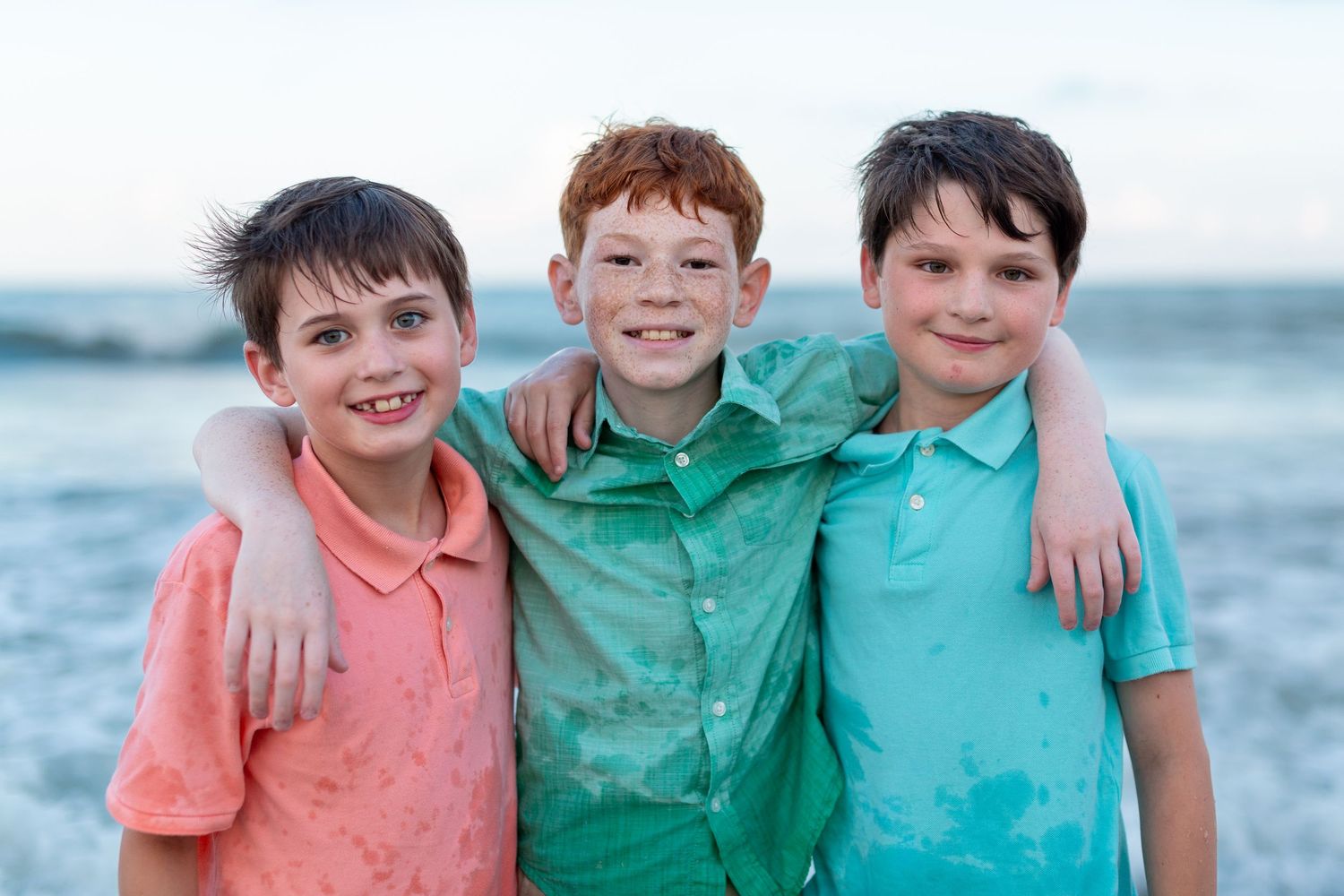 Three young boys in colorful shirts pose together smiling at the beach.