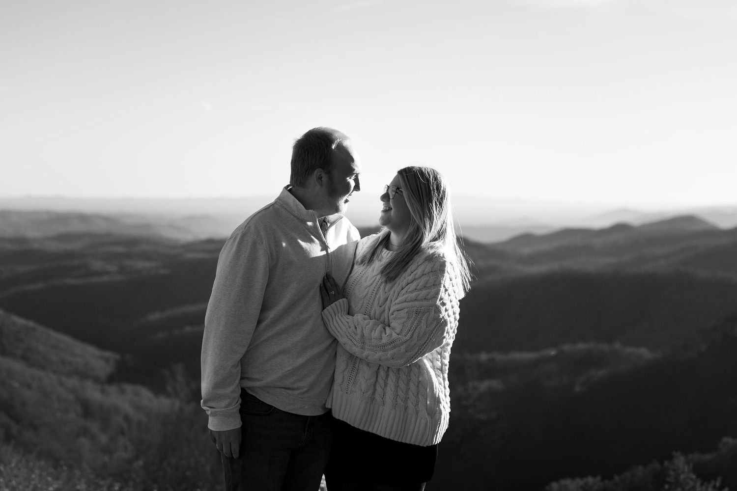 A couple embraces while overlooking a scenic mountain landscape at sunset in black and white.