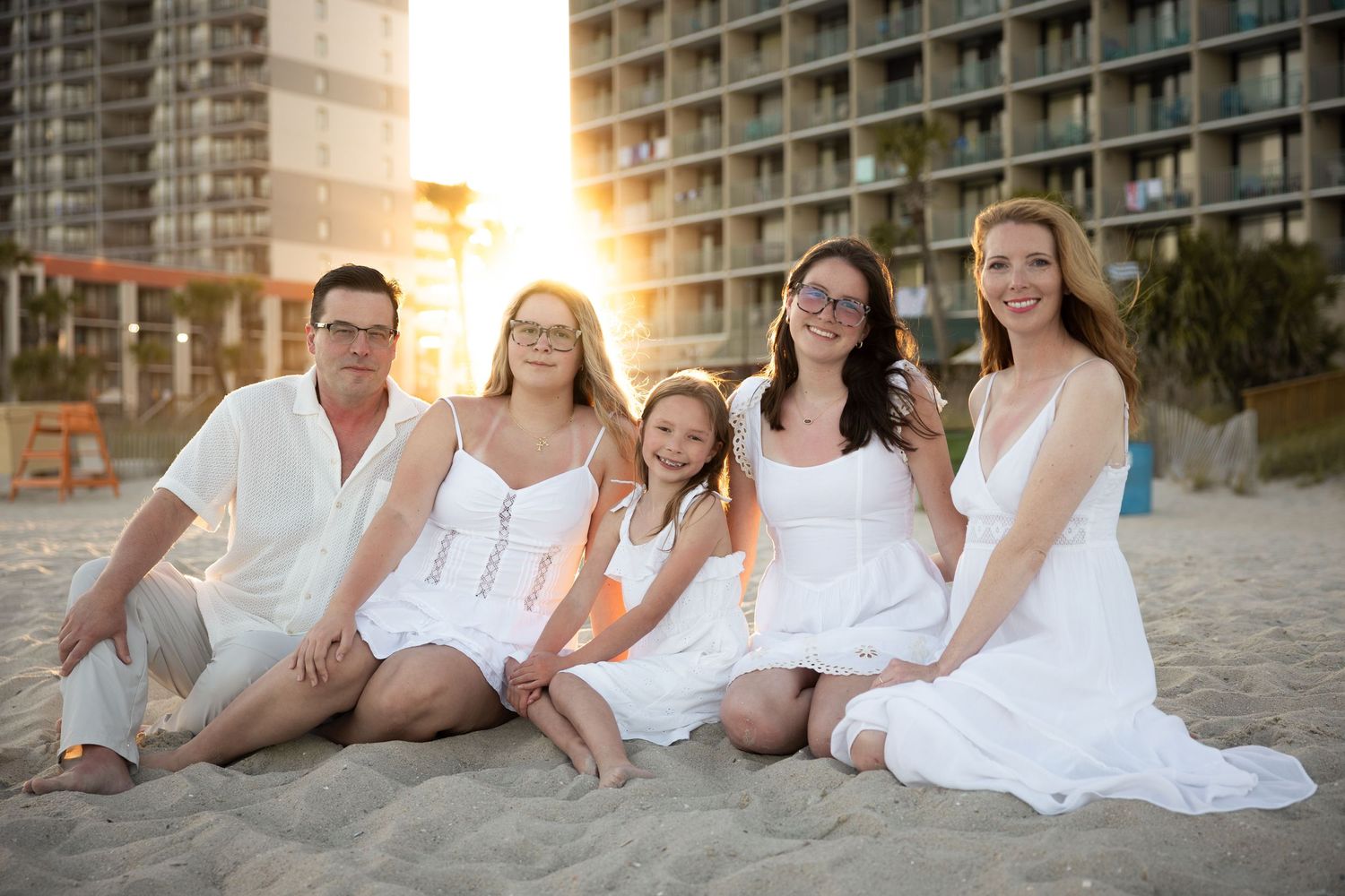 Family wearing white clothes posing together on sandy beach at sunset.