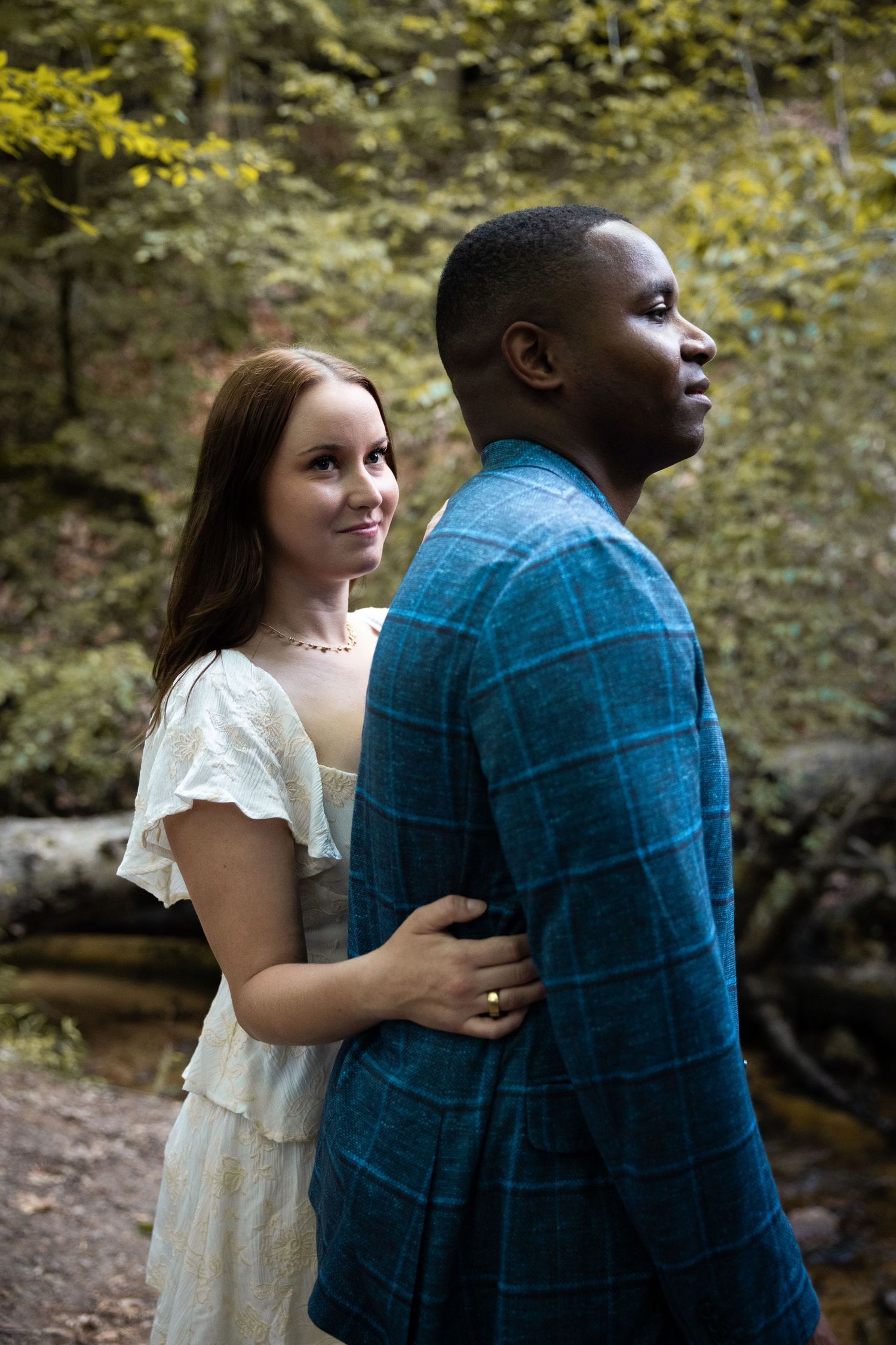 Couple in white dress and blue shirt embracing in lush forest setting.