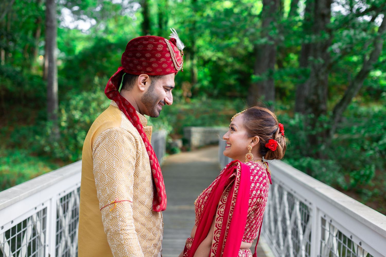 Couple in traditional Indian wedding attire smiling at each other on a white bridge surrounded by green trees.