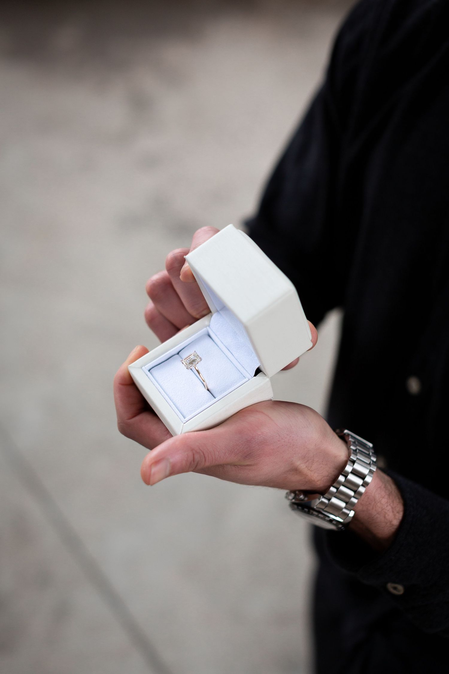 Someone holding a small white jewelry box with a ring inside while wearing a silver metal watch.