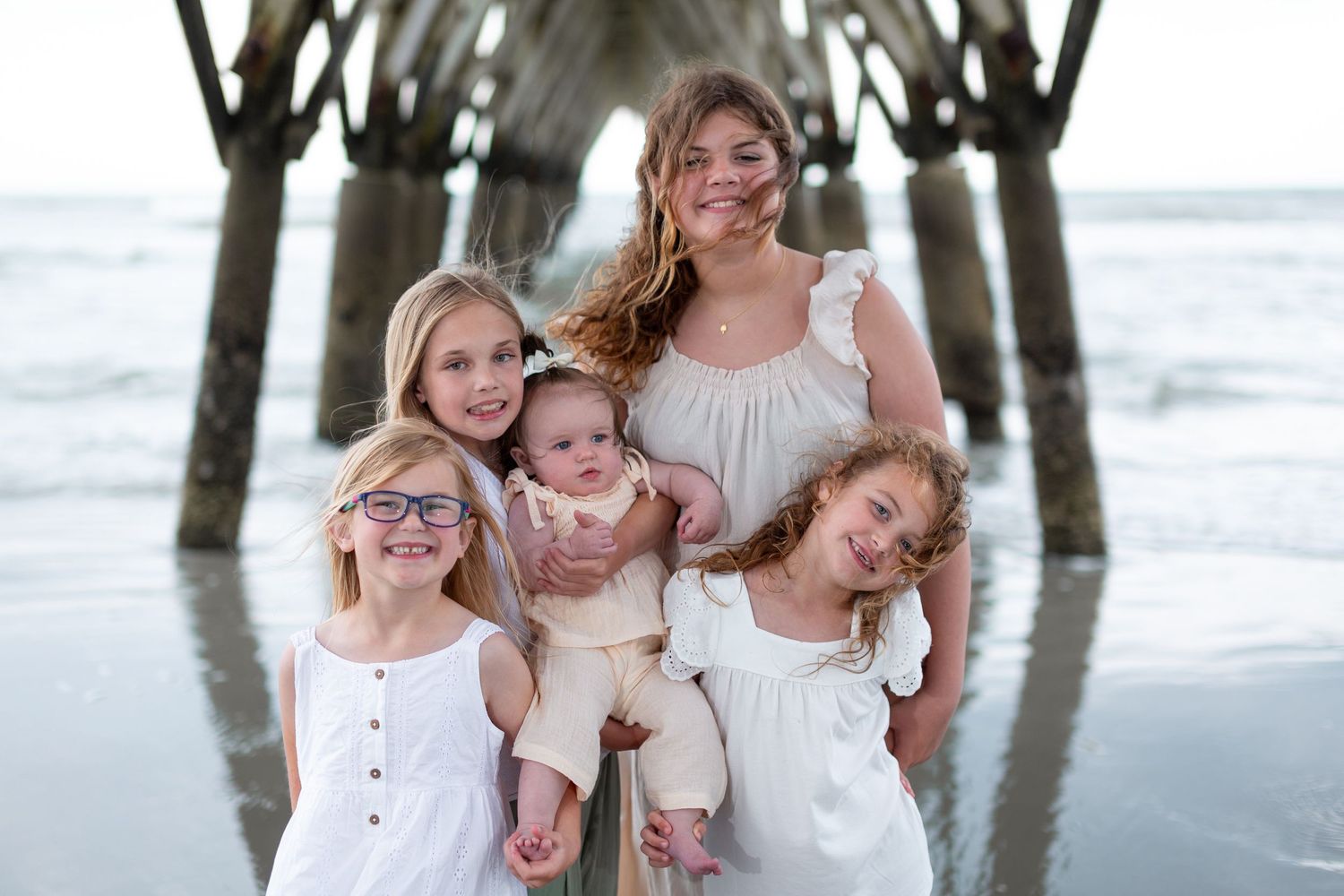 A family wearing coordinated white outfits poses together under a pier at the beach during sunset.