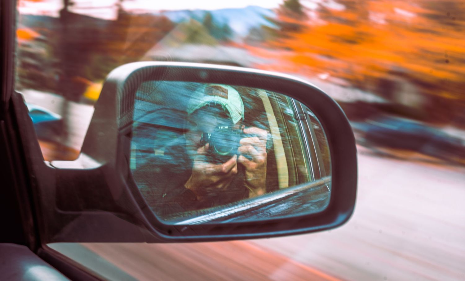 Person photographed in car side mirror while driving past colorful autumn trees at speed.