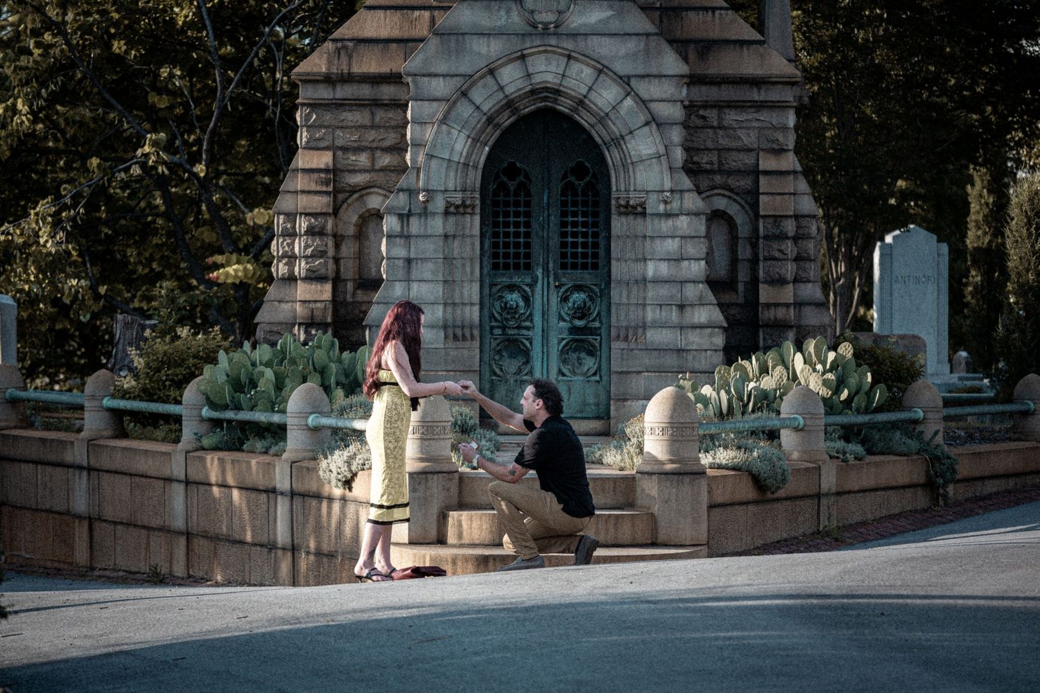 Man proposing to woman on one knee in front of ornate stone monument in a scenic outdoor setting.