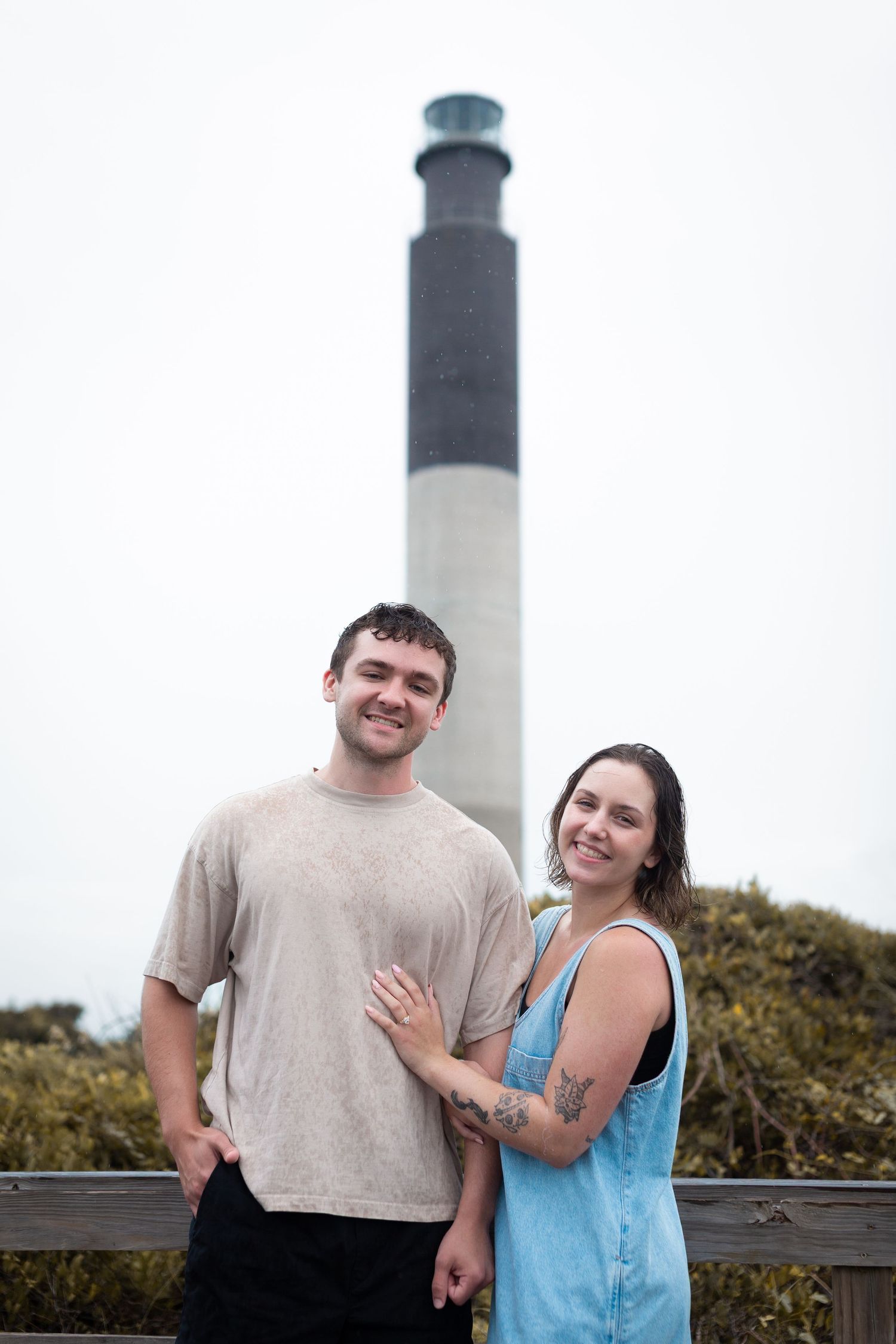 A couple stands together smiling in front of the Oak Island Lighthouse in North Carolina on a cloudy day.