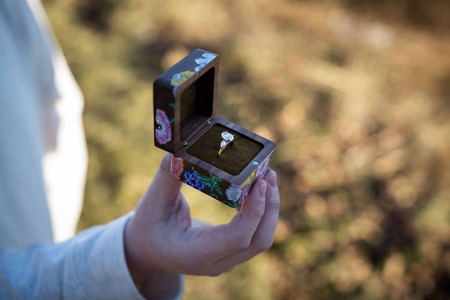 Person using smartphone outdoors while holding a small blue box in sunlit natural setting.