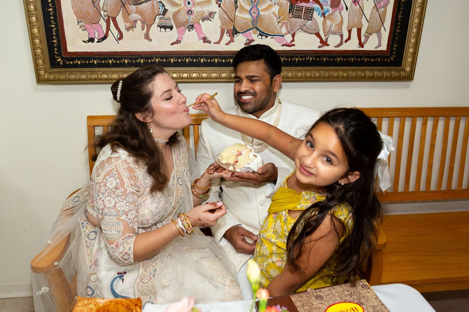 Family shares a joyful moment at home as a girl in a yellow dress feeds a sweet to her mother beside her father.