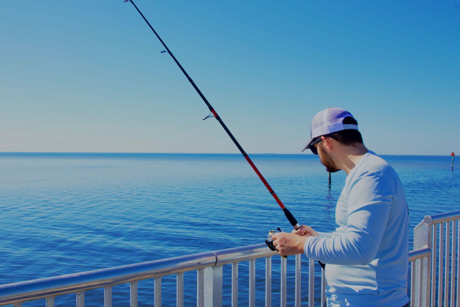 Man in white shirt and cap fishing from a pier over calm blue ocean waters on a clear sunny day.