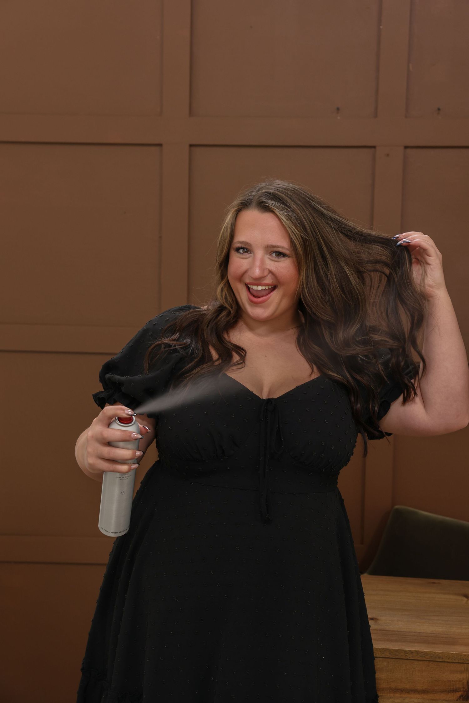 A smiling woman in a black dress sprays hairspray while holding her long brunette hair.