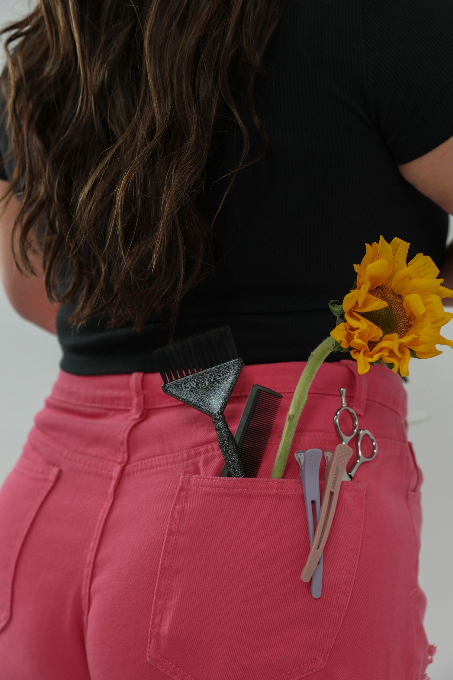 Close-up of pink jeans back pocket holding hair scissors, clips, comb, and a sunflower.