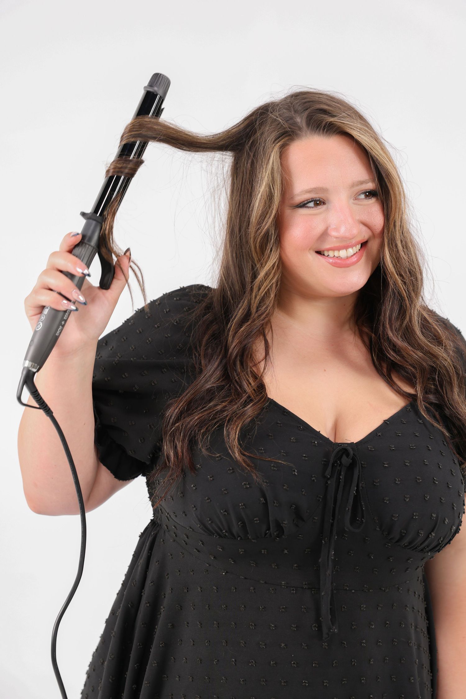 Smiling woman in black dress using a curling iron on her long wavy highlighted hair.