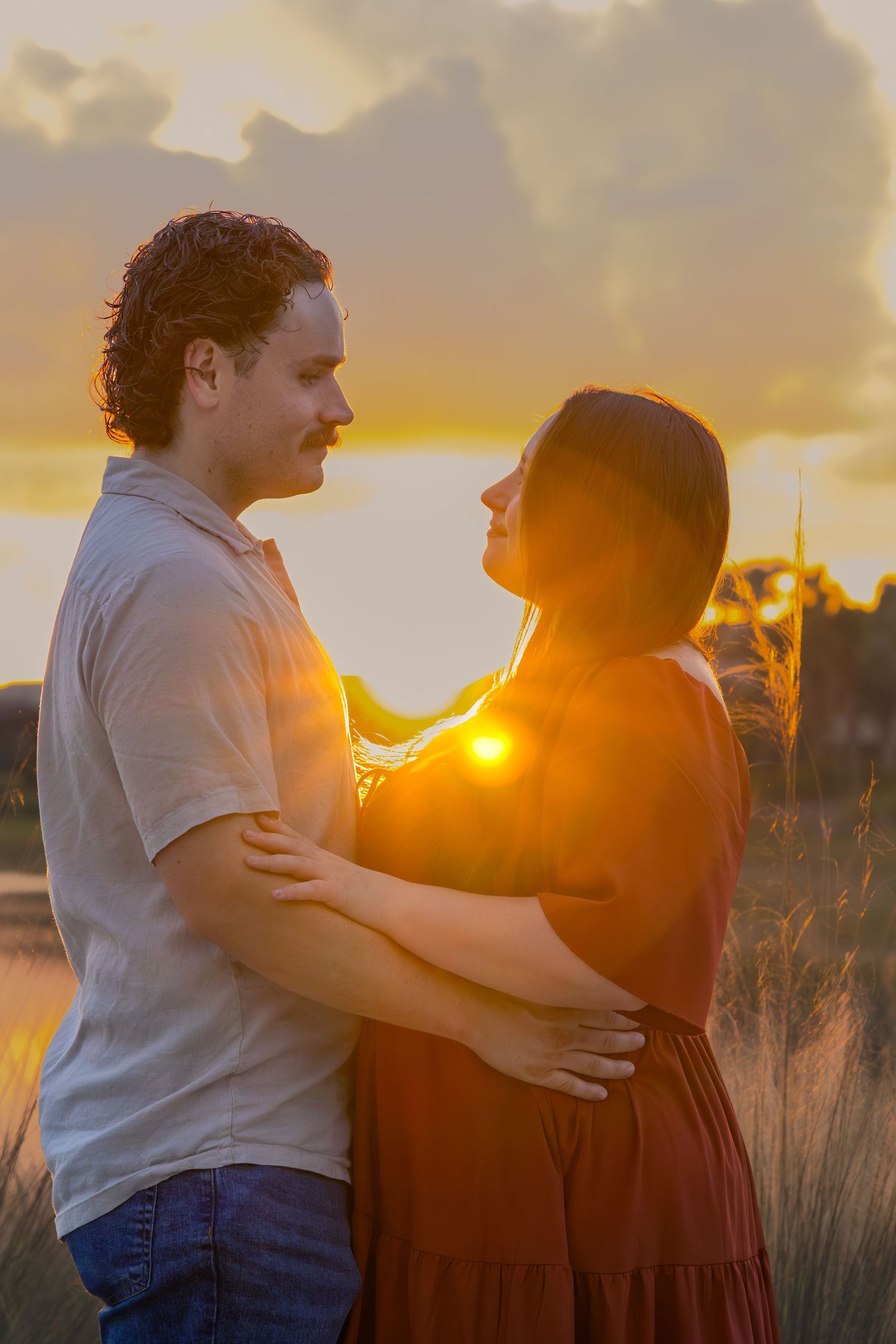 Couple embraces with glowing golden sunset bursting between them in a field.