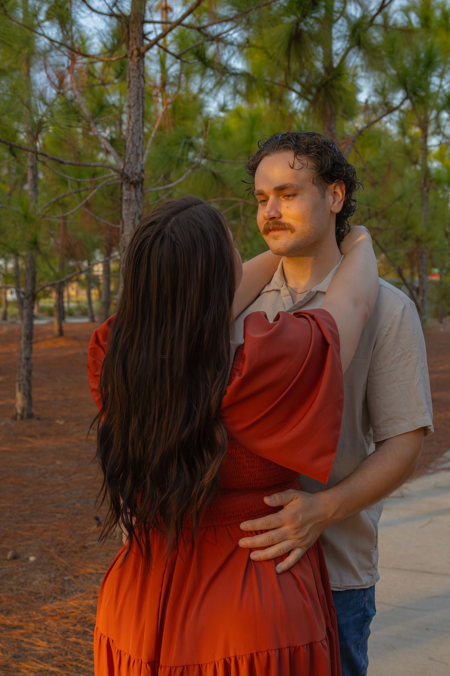 A couple embraces romantically at sunset in a pine forest, woman in red dress, man in beige shirt.