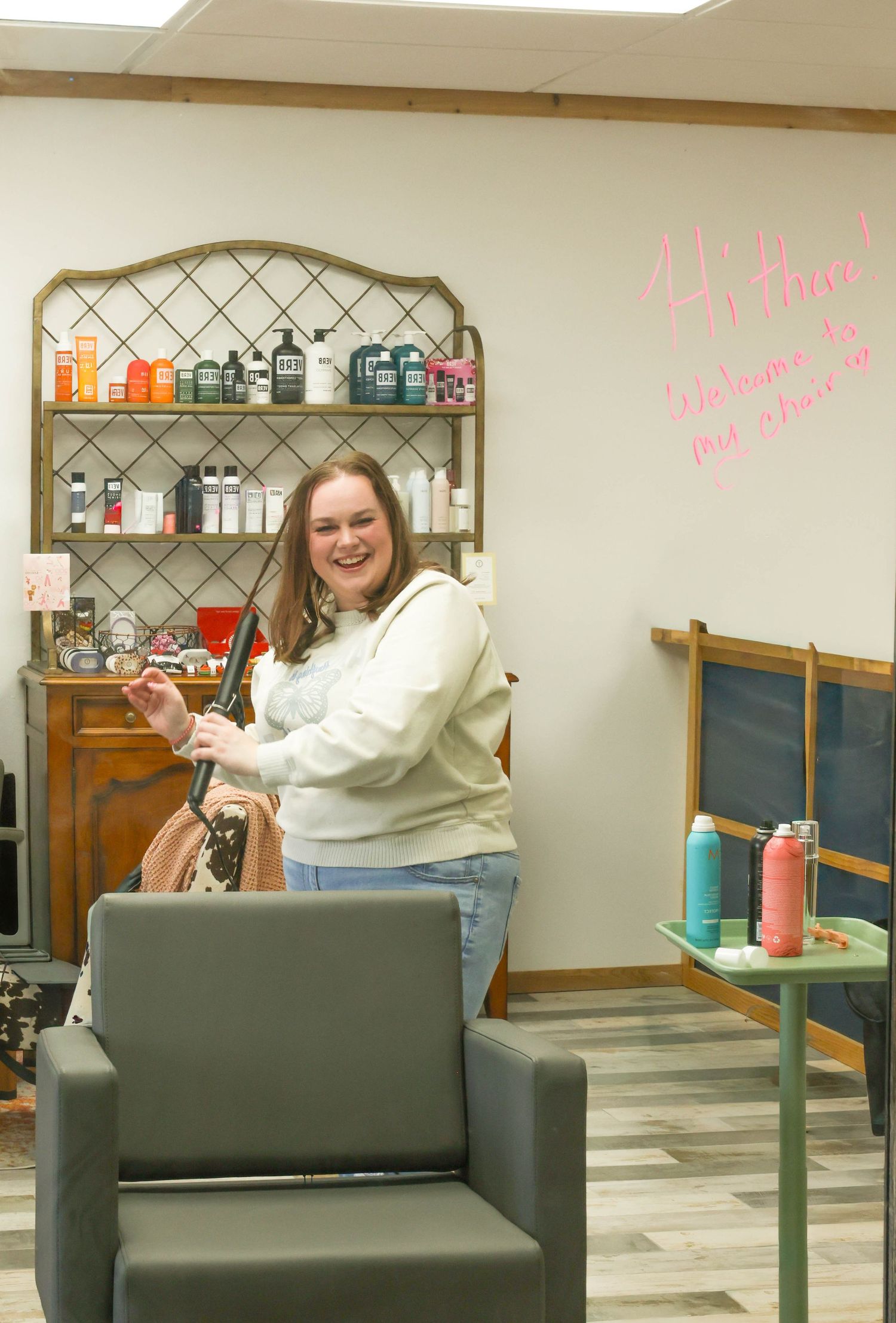 Smiling woman in cream sweatshirt standing in a hair salon with product shelves and welcome wall writing.