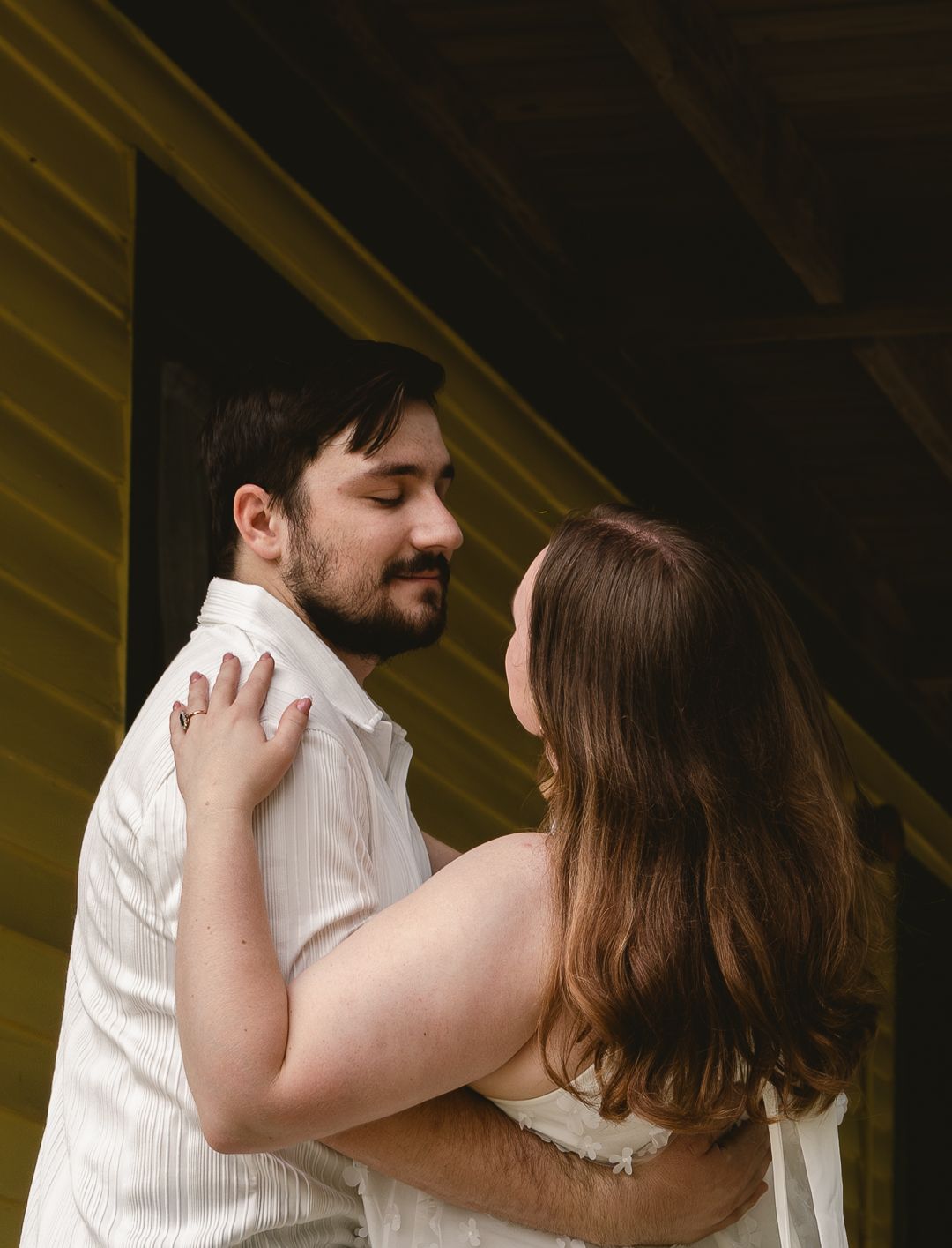A couple sharing a tender moment, the man in a white shirt smiling as the woman holds him close outdoors.