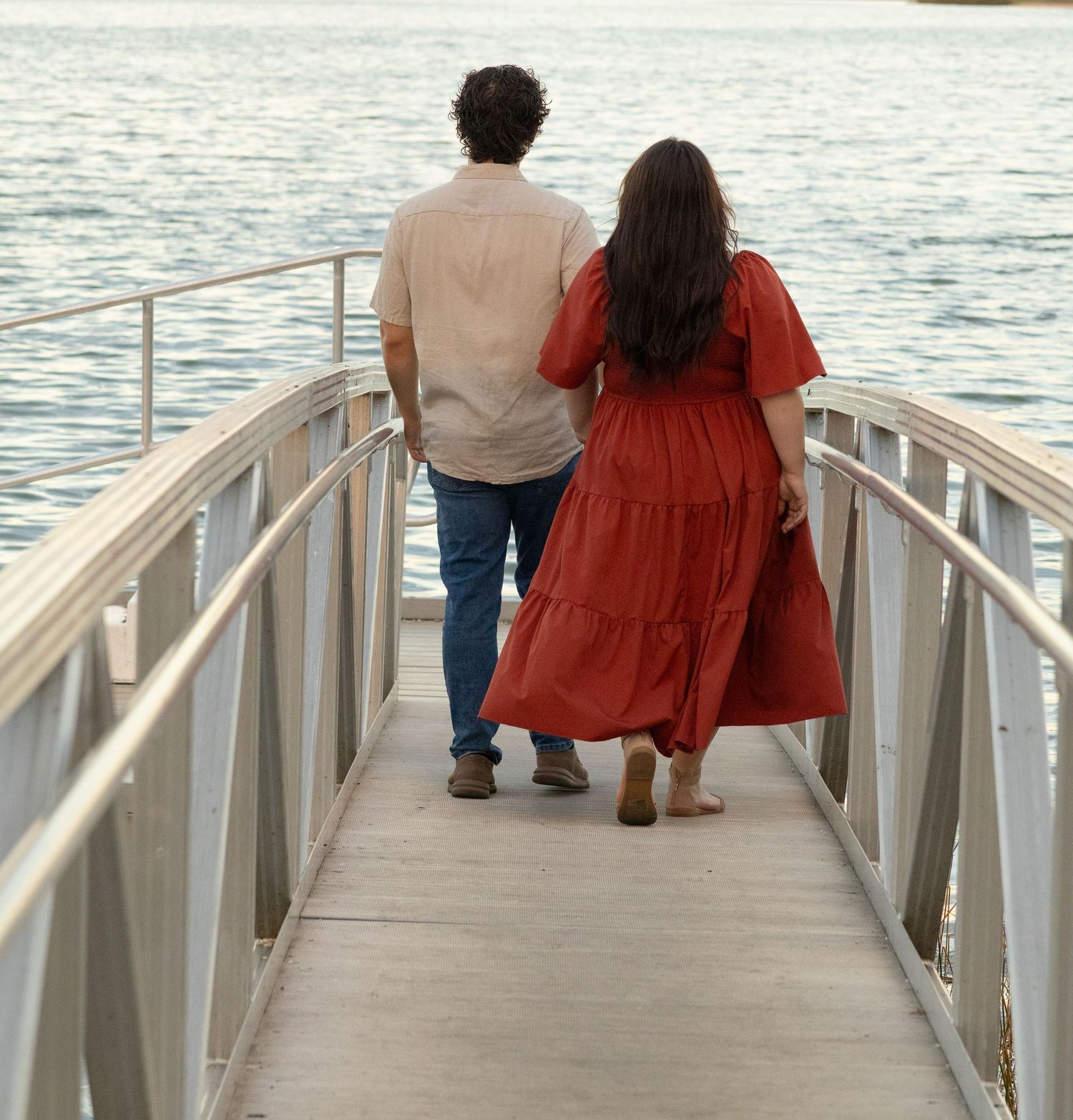 Couple walking hand in hand down a waterfront pier, woman in red dress and man in beige shirt and jeans.