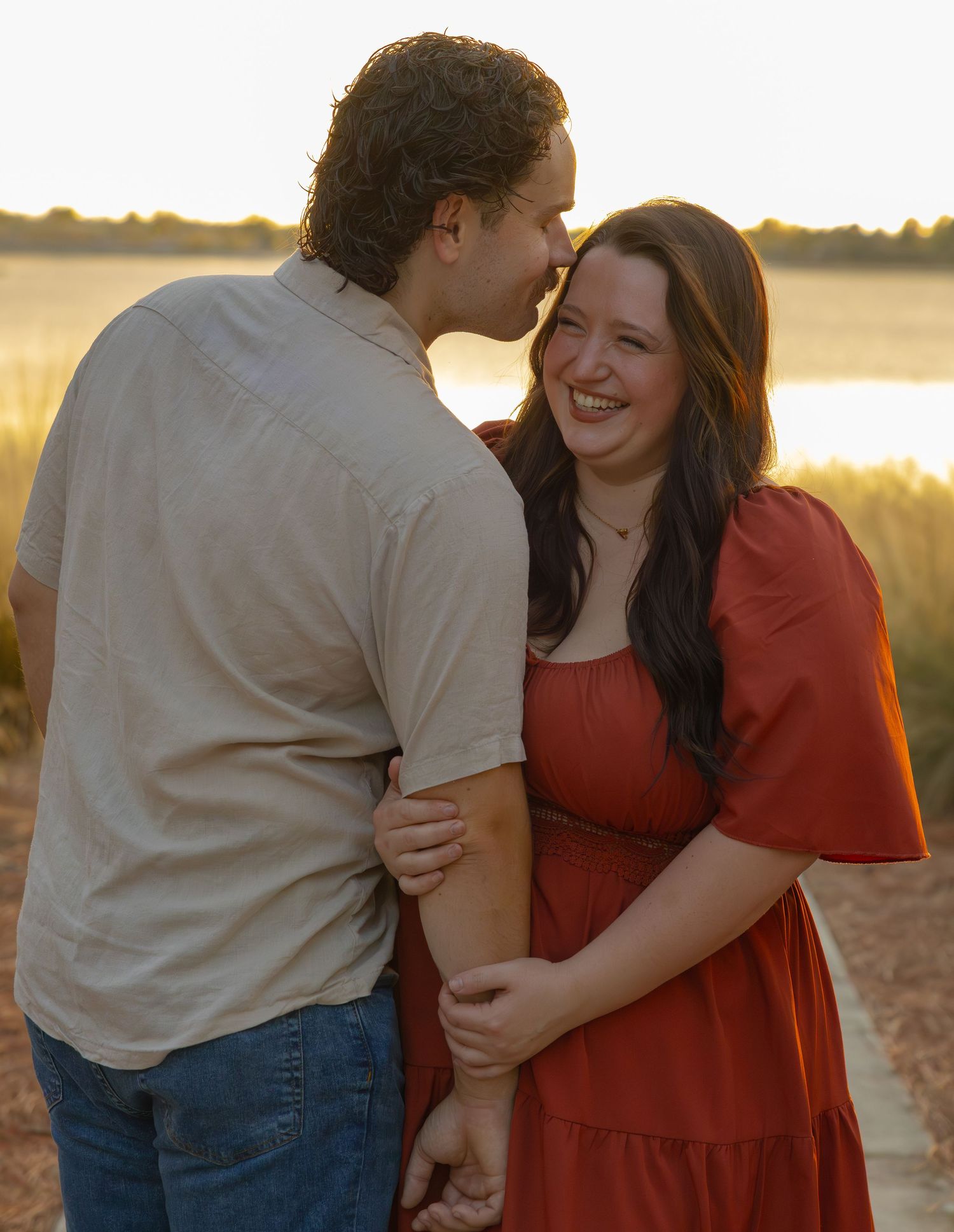 A joyful couple laughs together at golden hour near a scenic waterfront, woman in a red dress.