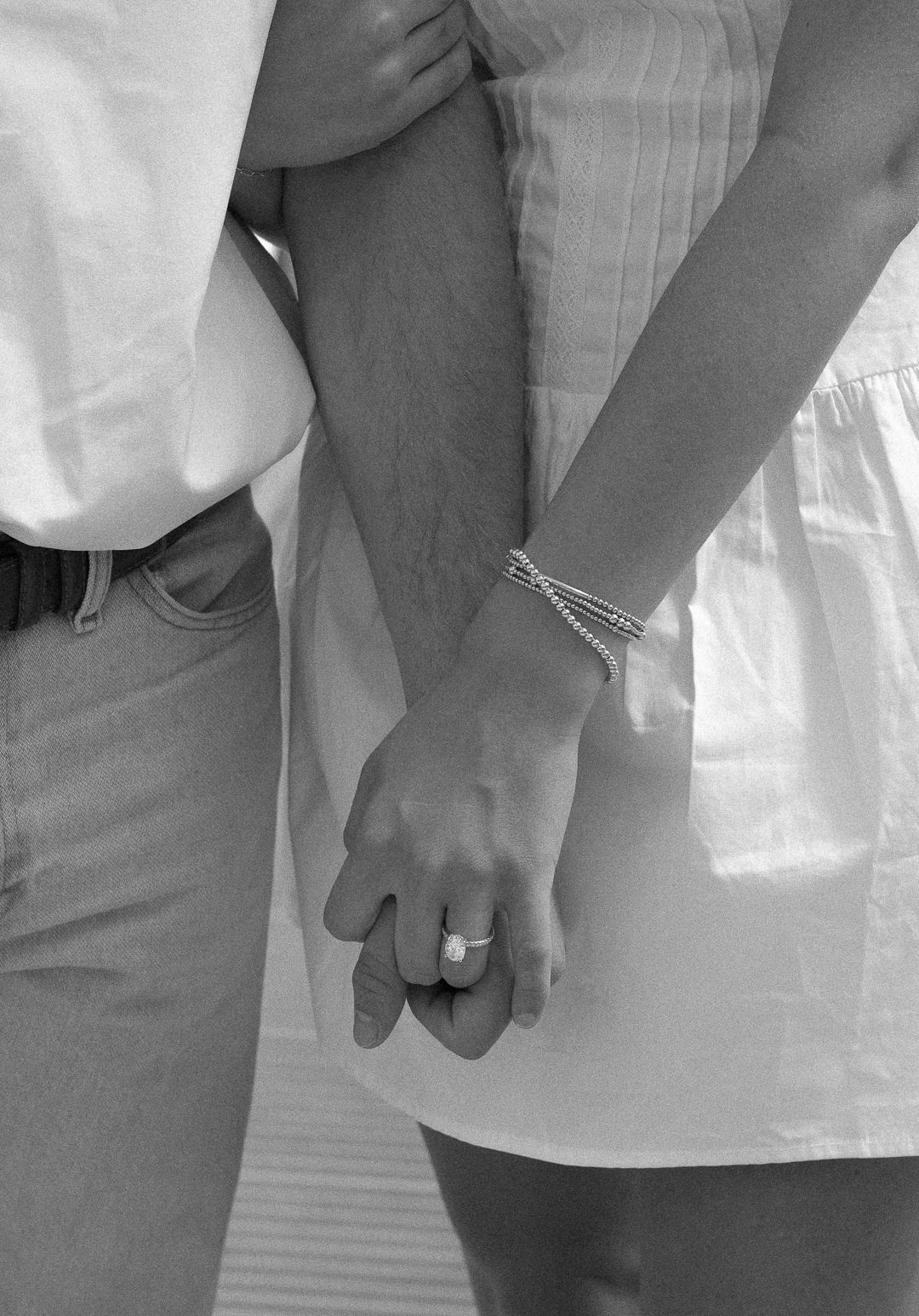 Black and white close-up of engaged couple holding hands, highlighting diamond engagement ring and delicate bracelets.